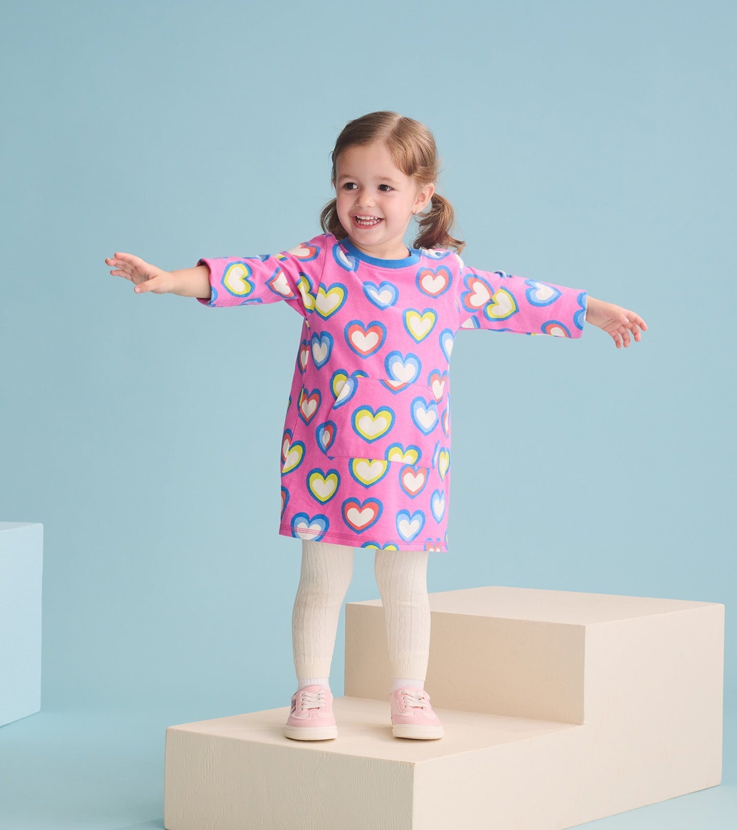 Pink terry dress with colorful heart pattern, long sleeves, and a relaxed fit, worn by a smiling girl on a pedestal.