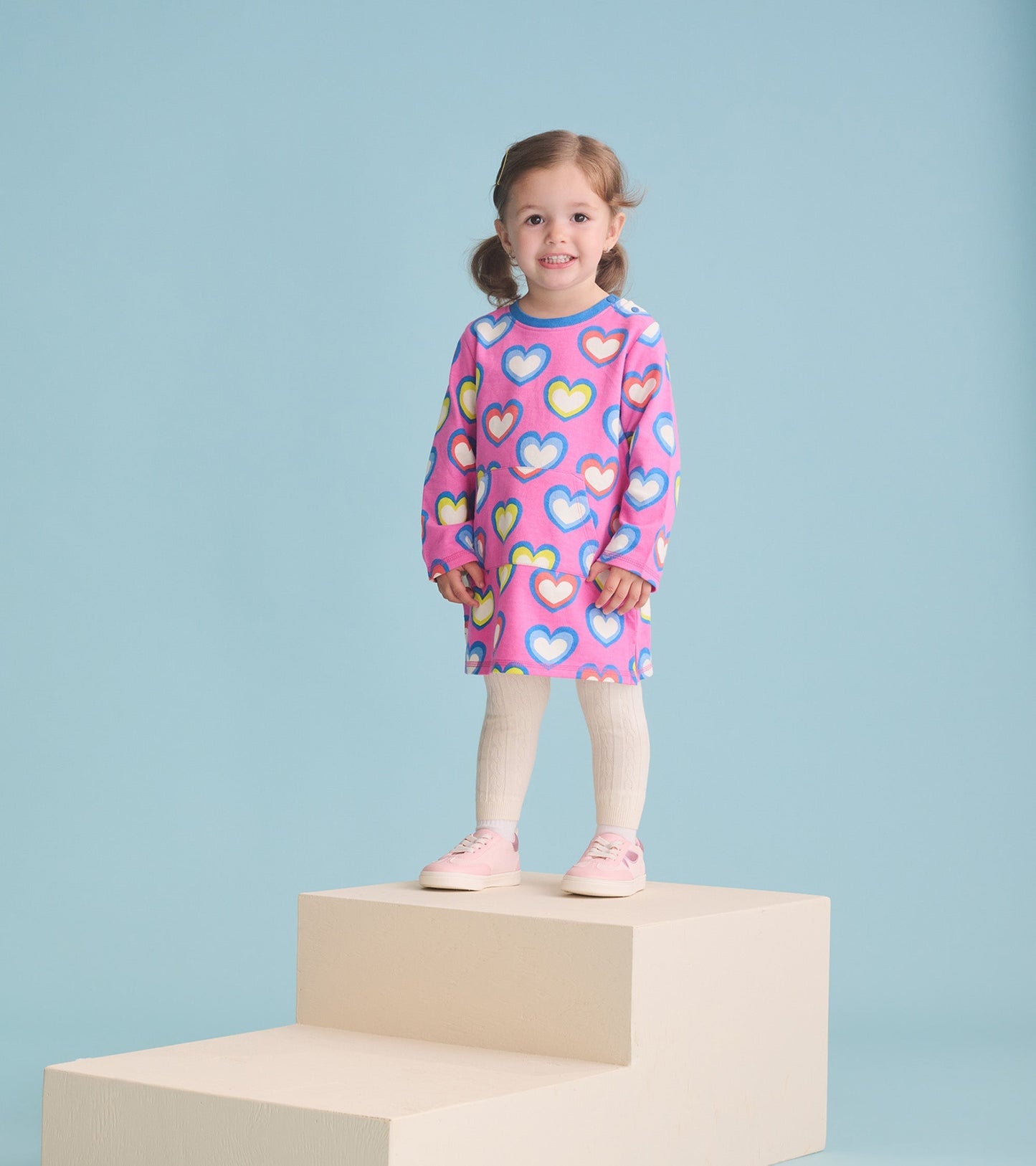 Pink terry dress with colorful heart patterns, long sleeves, and a relaxed fit, worn by a girl standing on a platform.