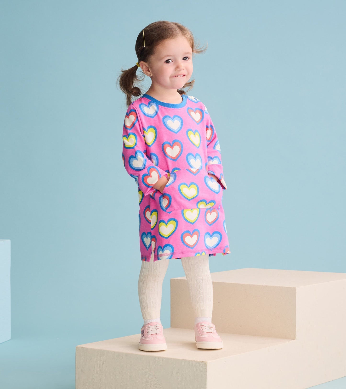 Pink terry dress with colorful heart pattern, long sleeves, and pockets, worn with white tights and pink shoes.