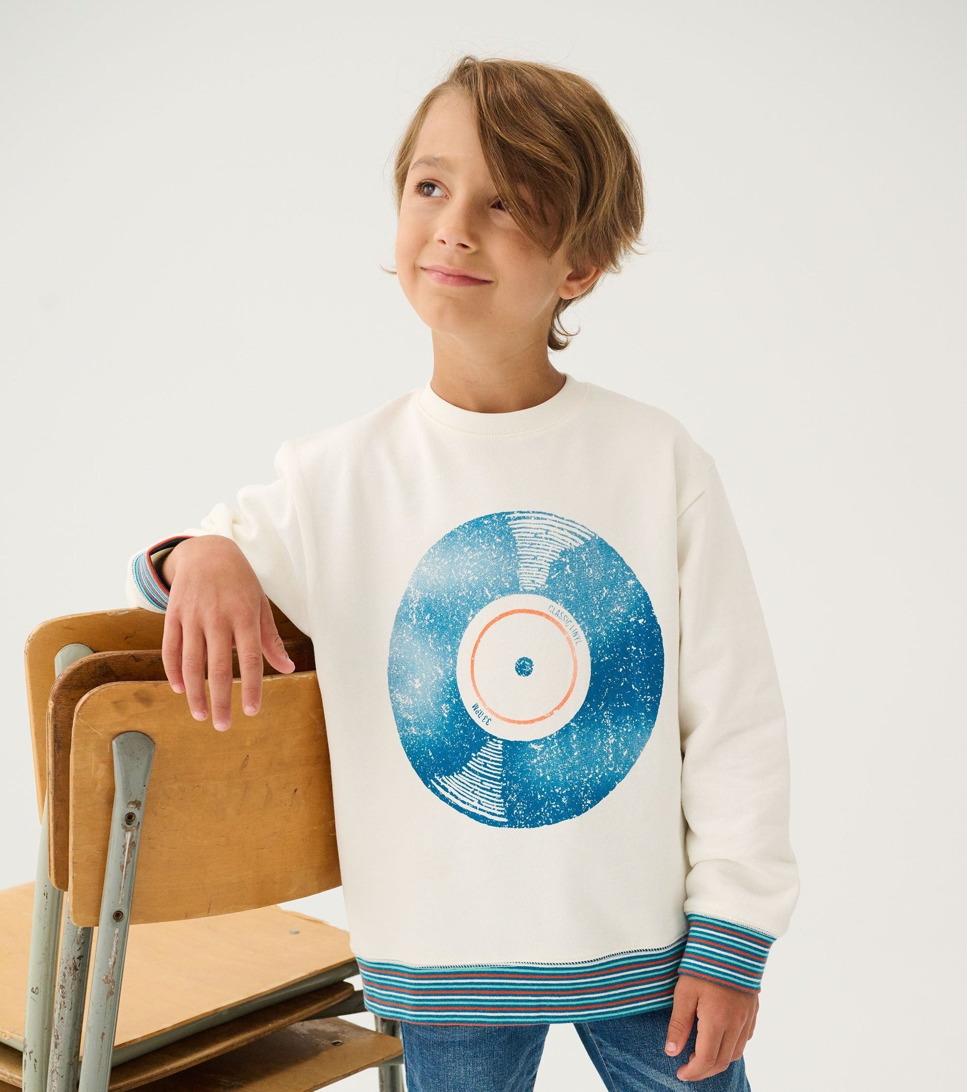 White sweatshirt featuring a blue vinyl record graphic and striped cuffs, worn by a boy leaning on a wooden chair.