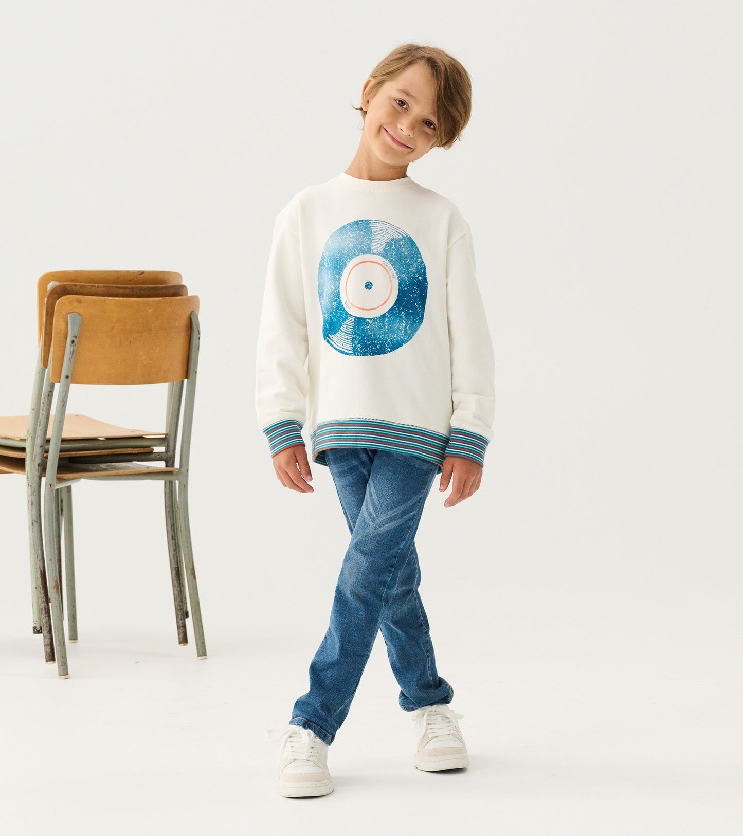White sweatshirt with a blue vinyl record graphic and striped cuffs, worn by a smiling boy with jeans and sneakers.