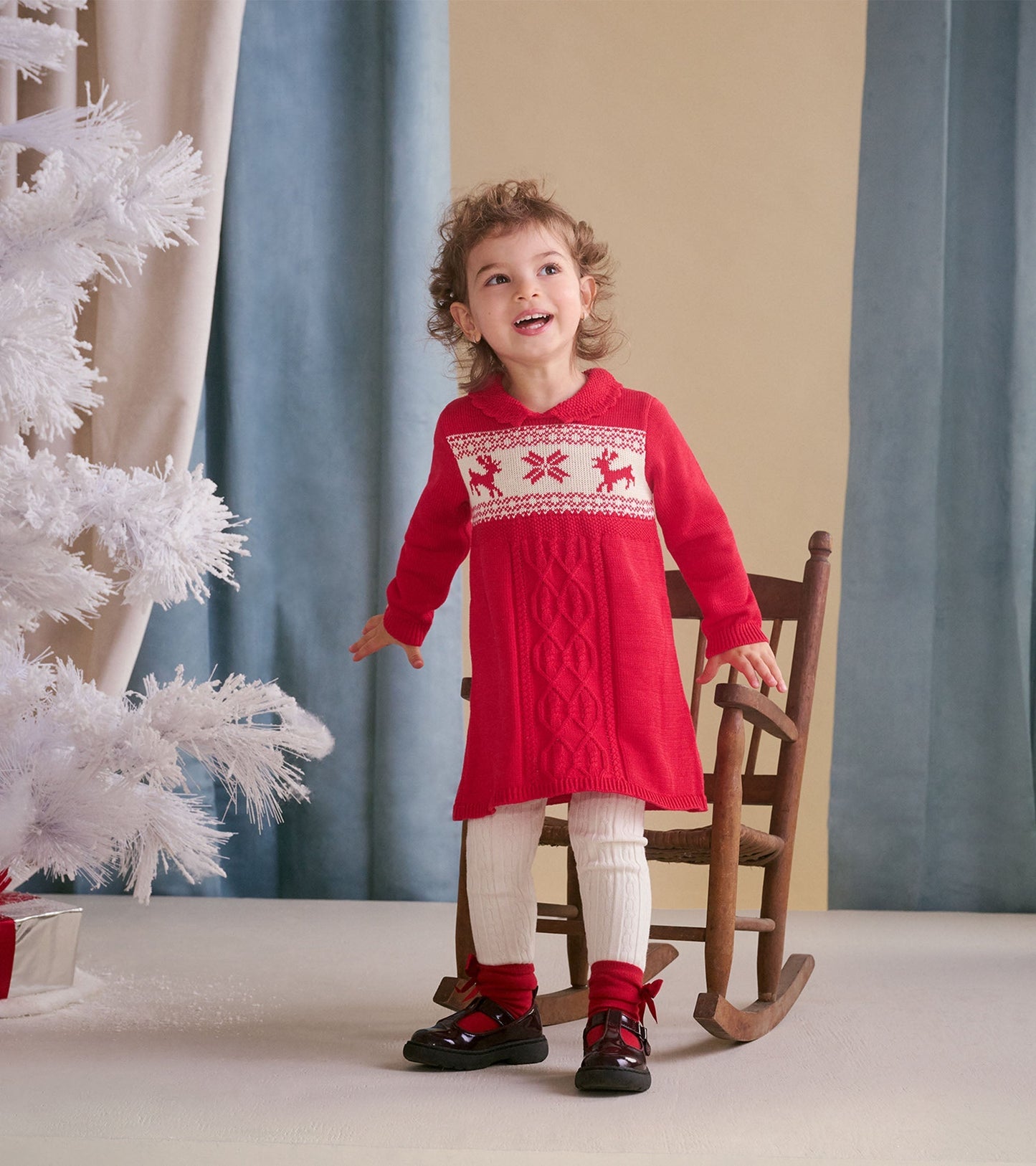 Red cable knit sweater dress with festive reindeer pattern, paired with white leggings and black shoes.