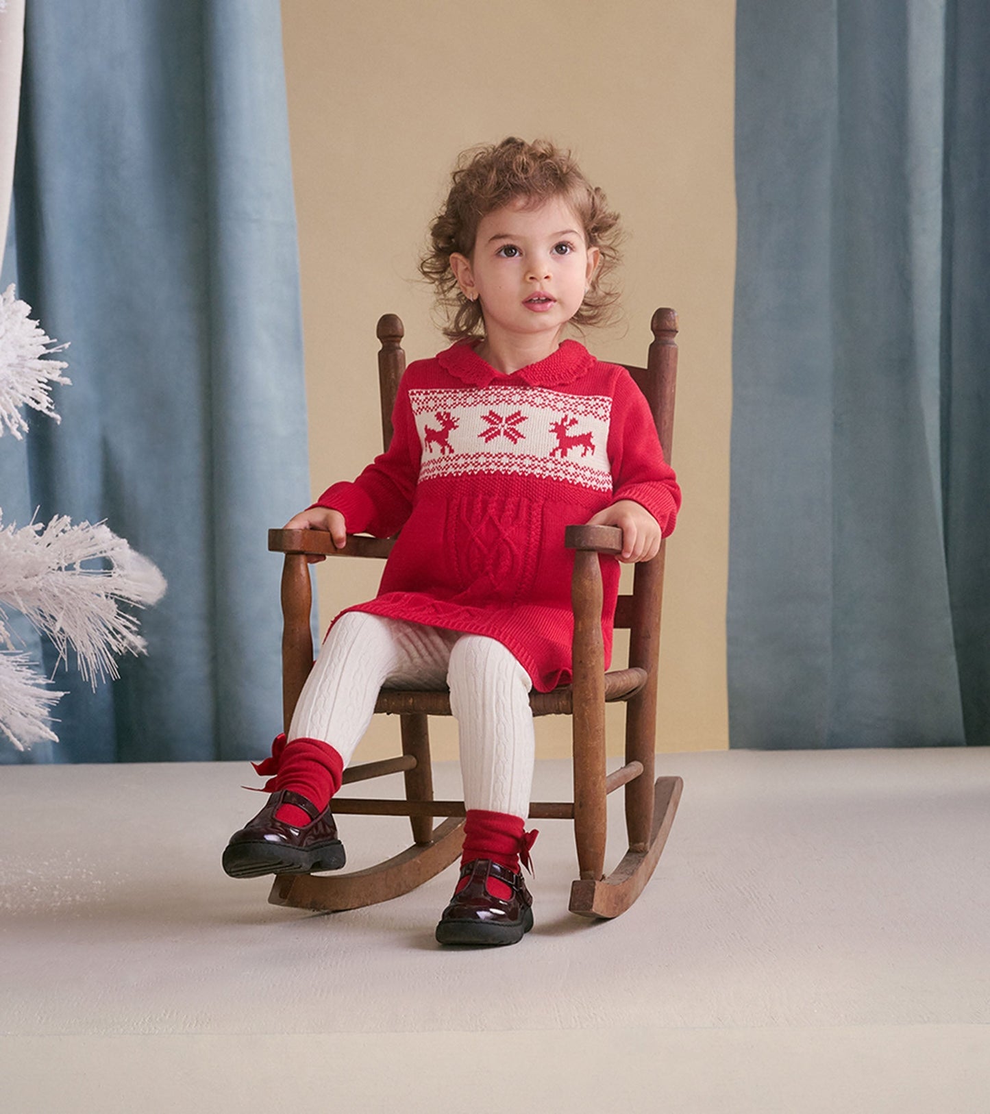 Red cable knit sweater dress with festive reindeer pattern, paired with white tights and black shoes, worn by a toddler.