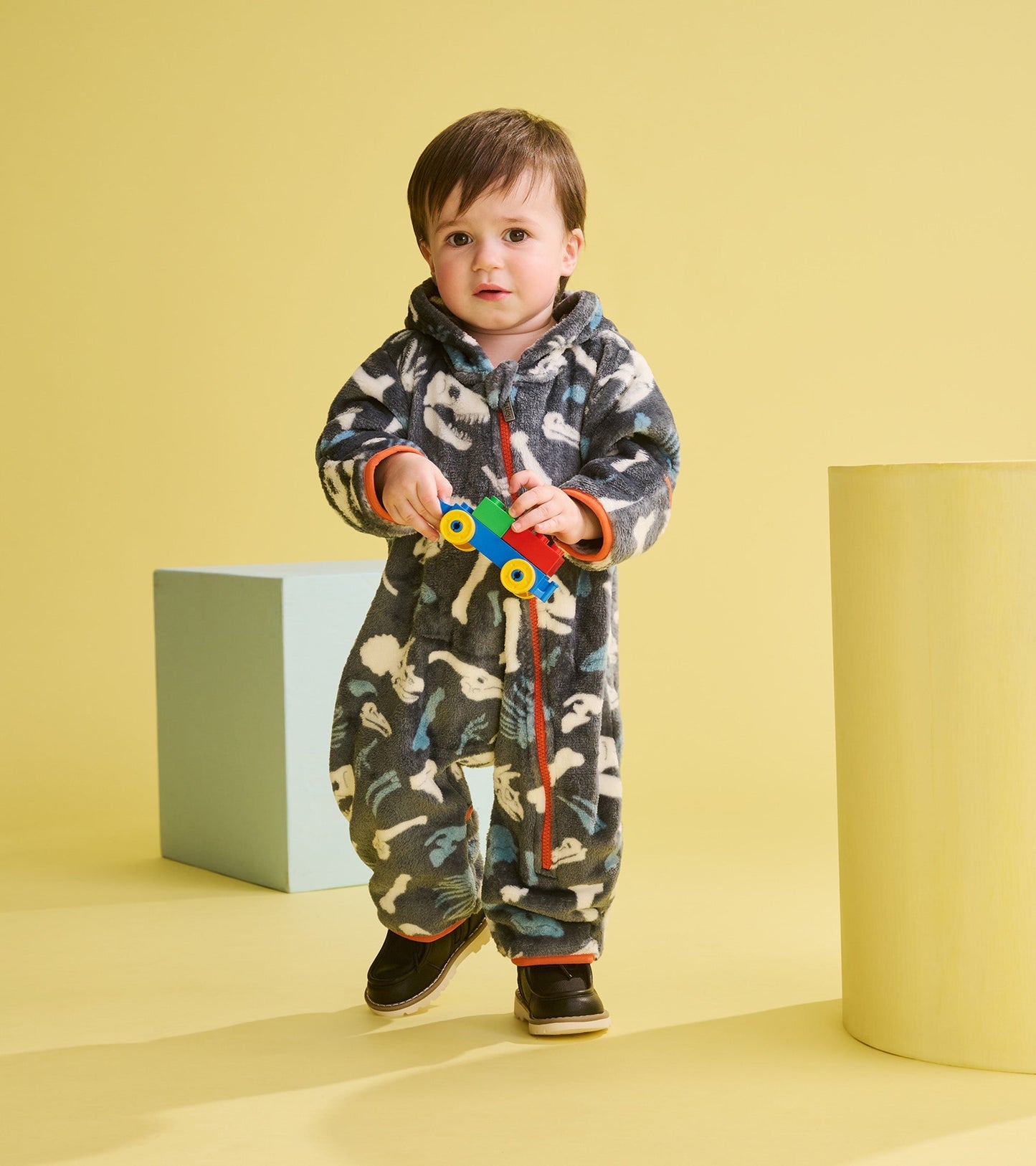 Gray fleece suit with dinosaur fossil pattern, featuring a hood and orange accents, worn by a toddler holding a toy.
