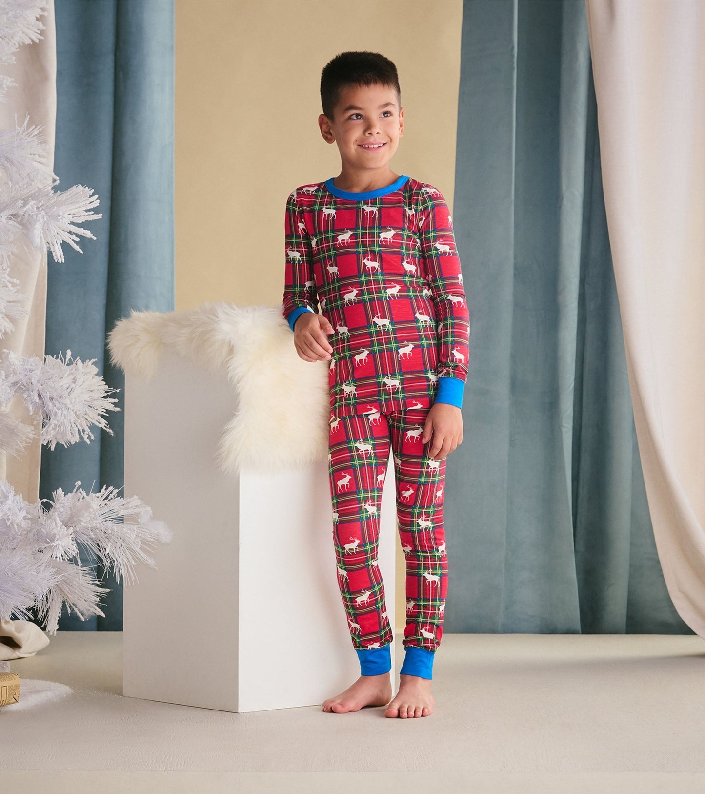 Red plaid pyjama set with blue cuffs, featuring elk patterns, worn by a smiling boy standing beside a white pedestal.