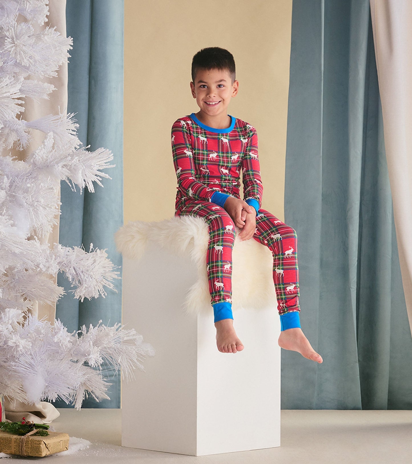 Red plaid pyjama set with elk print, featuring a blue collar and cuffs, worn by a smiling boy sitting on a white pedestal.