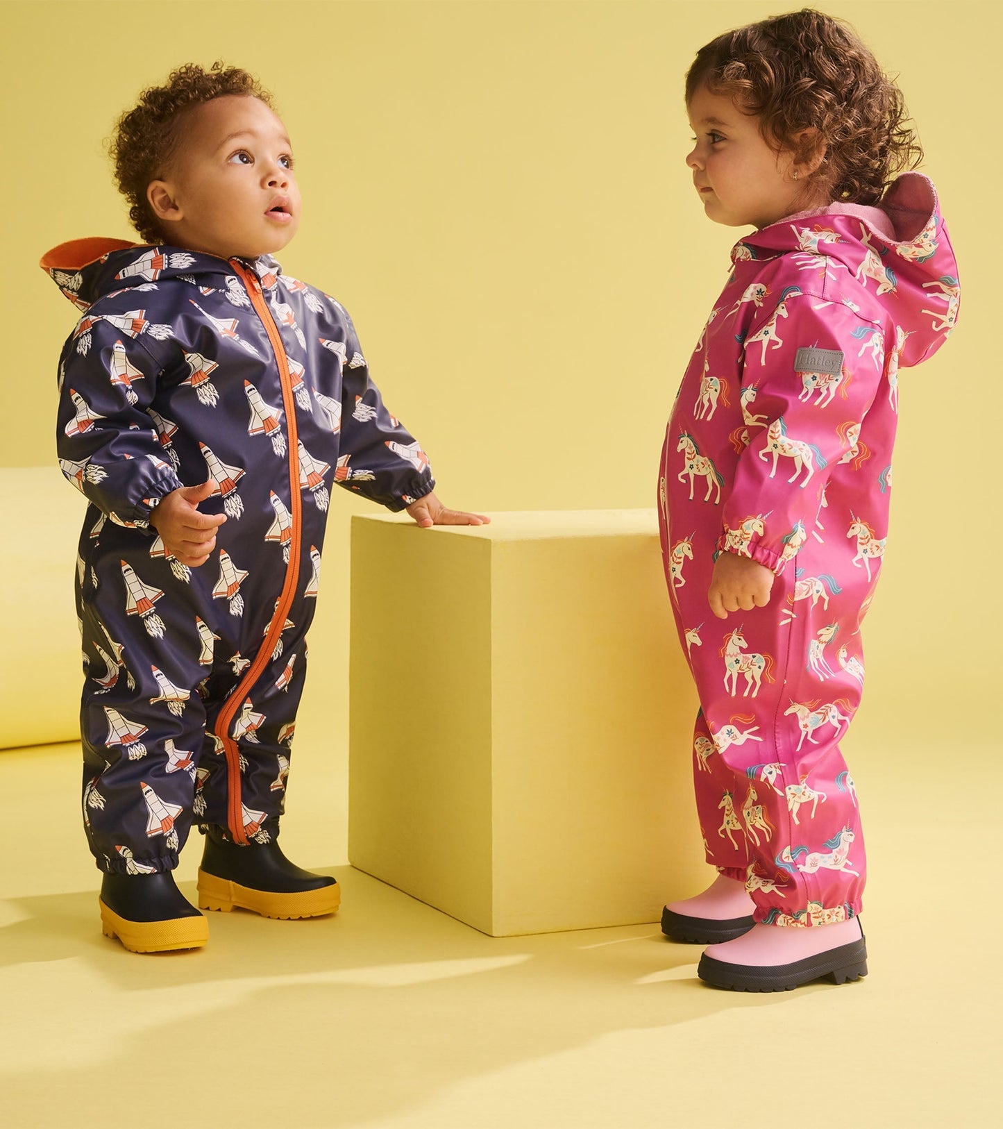 Two toddlers in colorful rain suits: one navy with rocket pattern, the other pink with unicorns, standing on a yellow backgr…