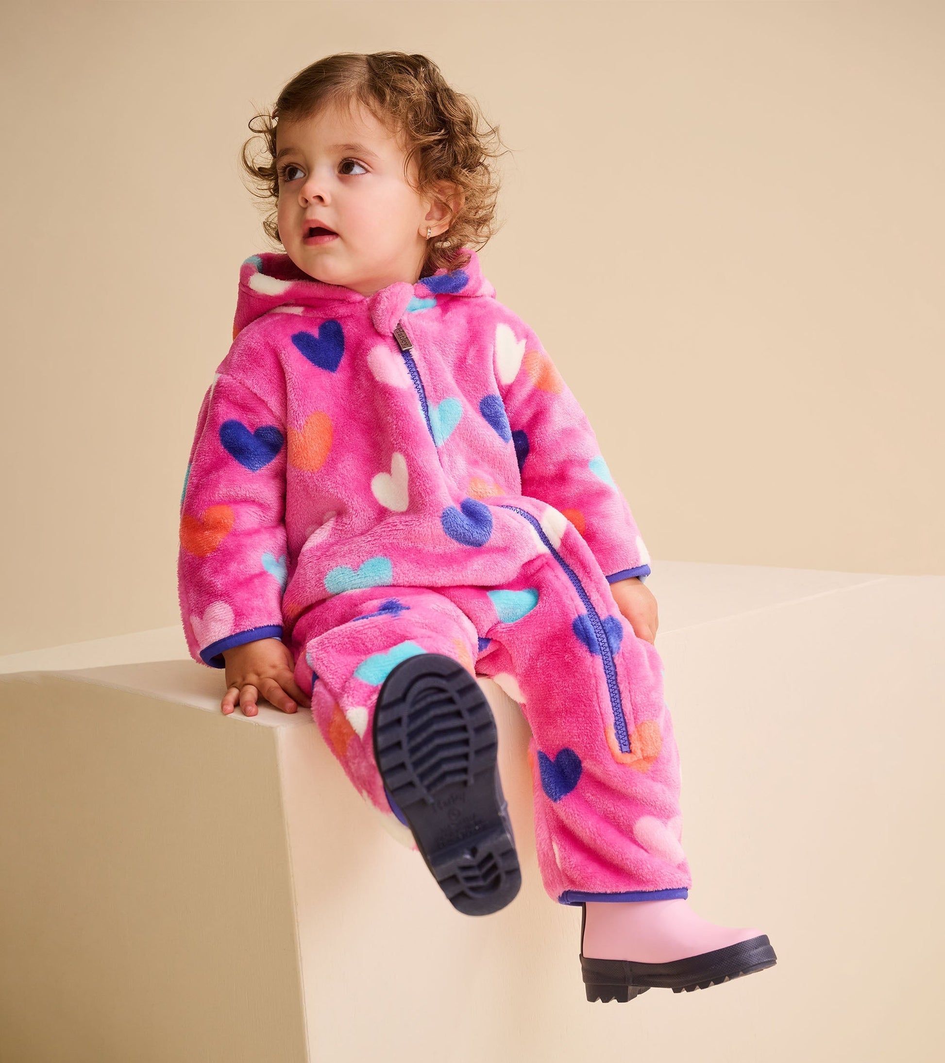 Pink fleece suit with colorful heart patterns, featuring a hood and zip closure, worn by a toddler sitting on a ledge.