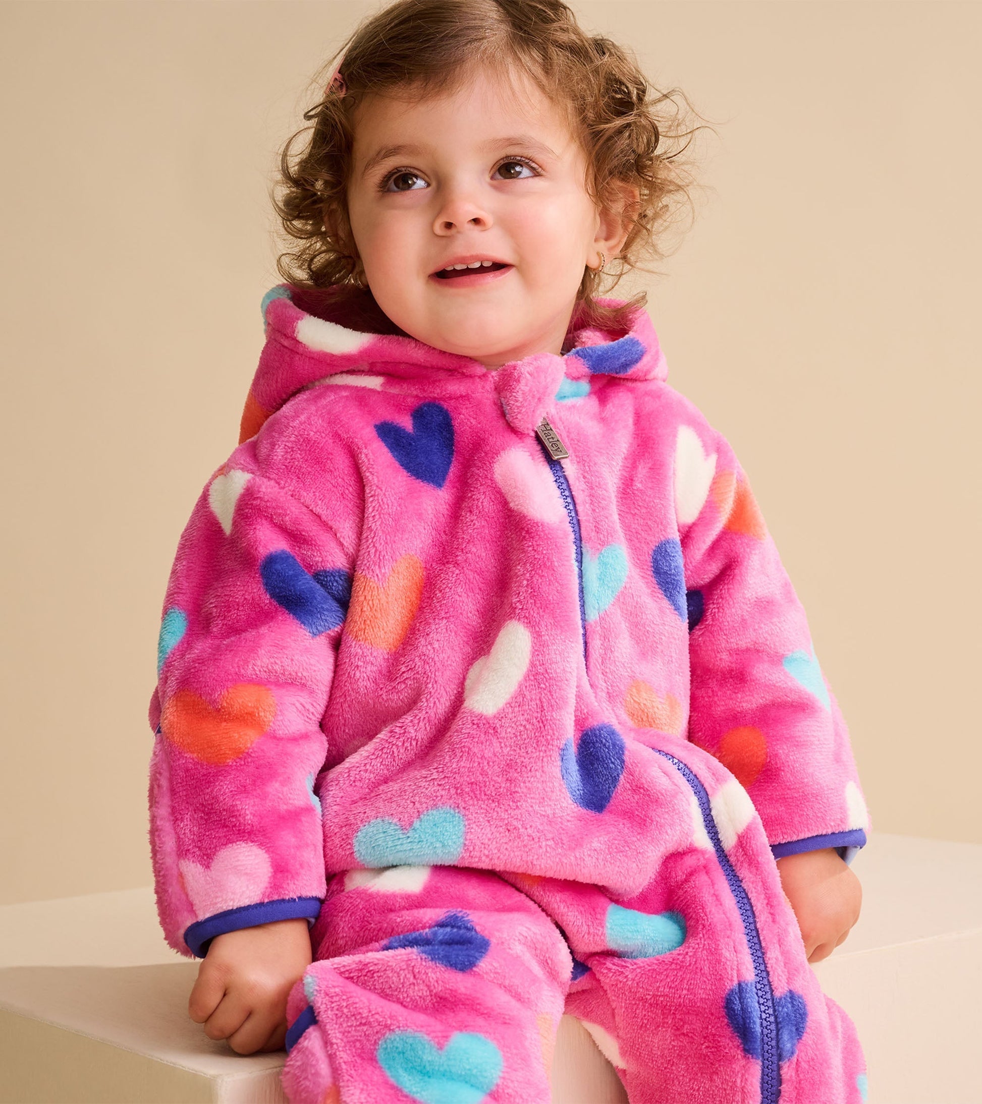 Pink fleece suit for baby girls featuring colorful heart patterns, with a zip front and hood. Suitable for cooler weather.