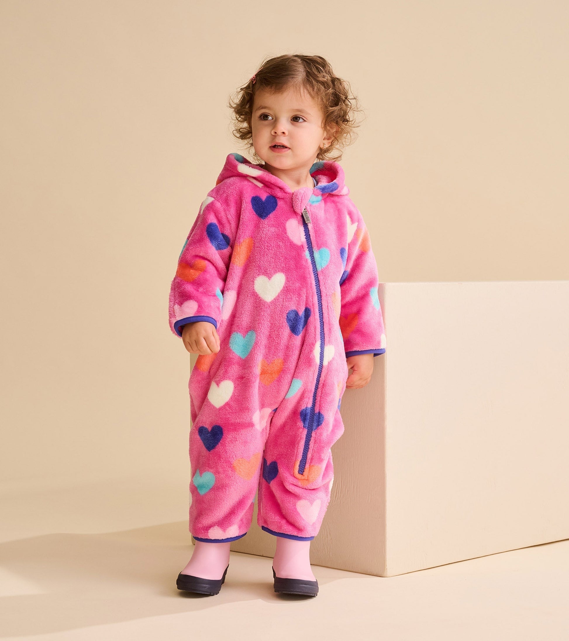 Pink fleece suit with colorful heart patterns, featuring a hood and zip closure, worn by a toddler.