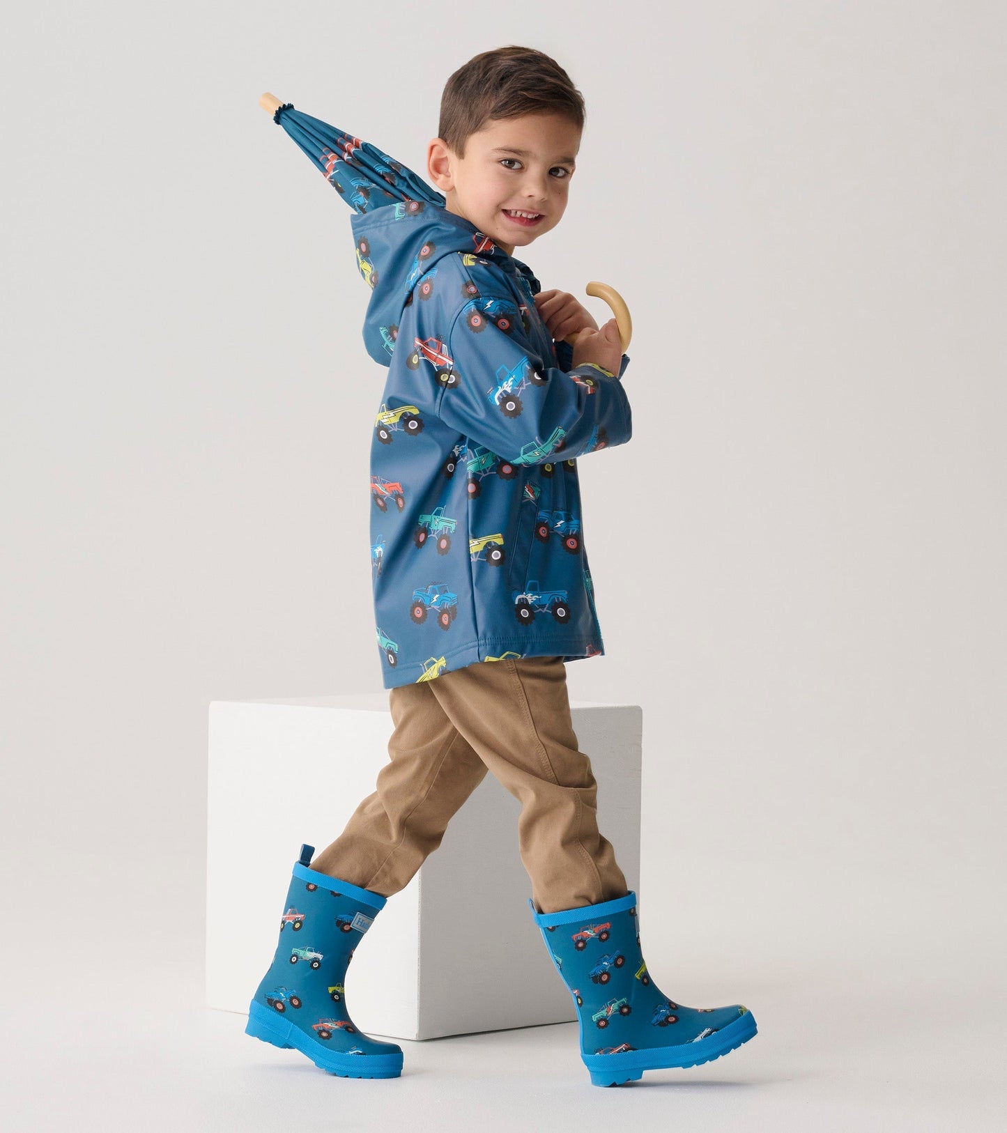 Blue matte wellies with colorful monster truck patterns, worn by a boy in a raincoat, standing beside a white block.