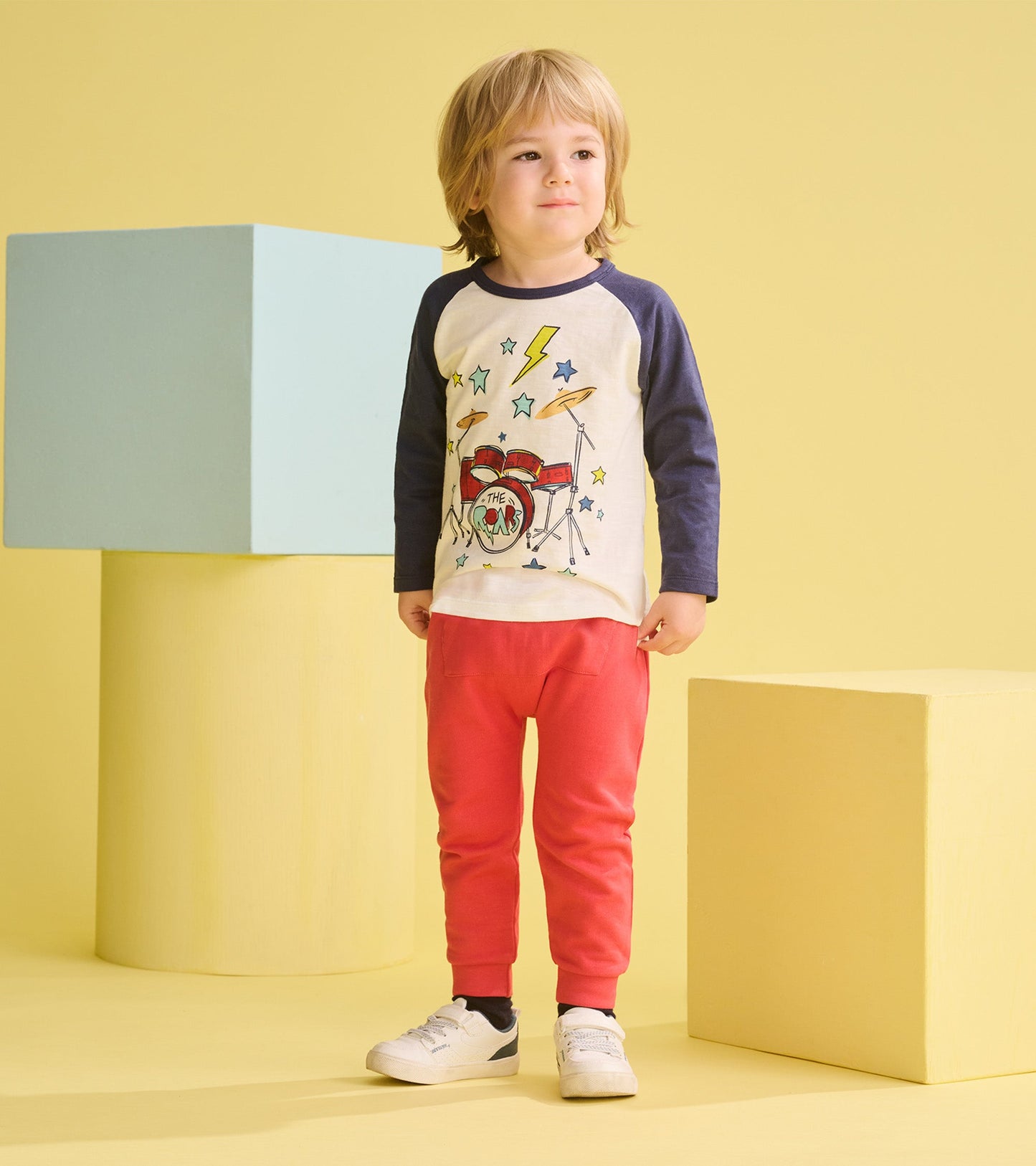 Long sleeve t-shirt featuring a drum kit graphic, paired with red pants, worn by a child against a yellow background.