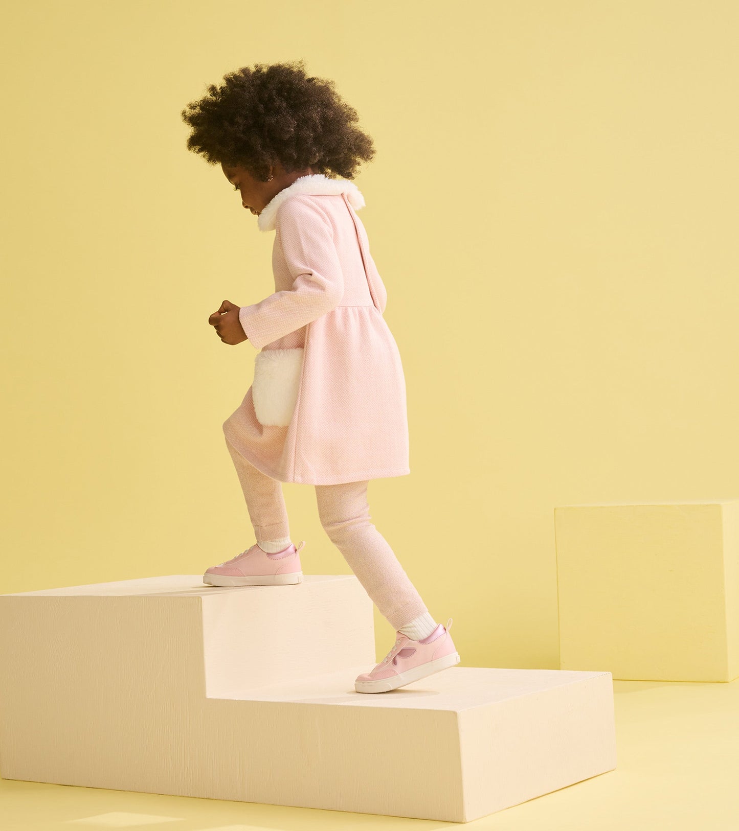 Pink herringbone dress with fur trim, paired with pink sneakers, as a toddler climbs steps against a yellow background.