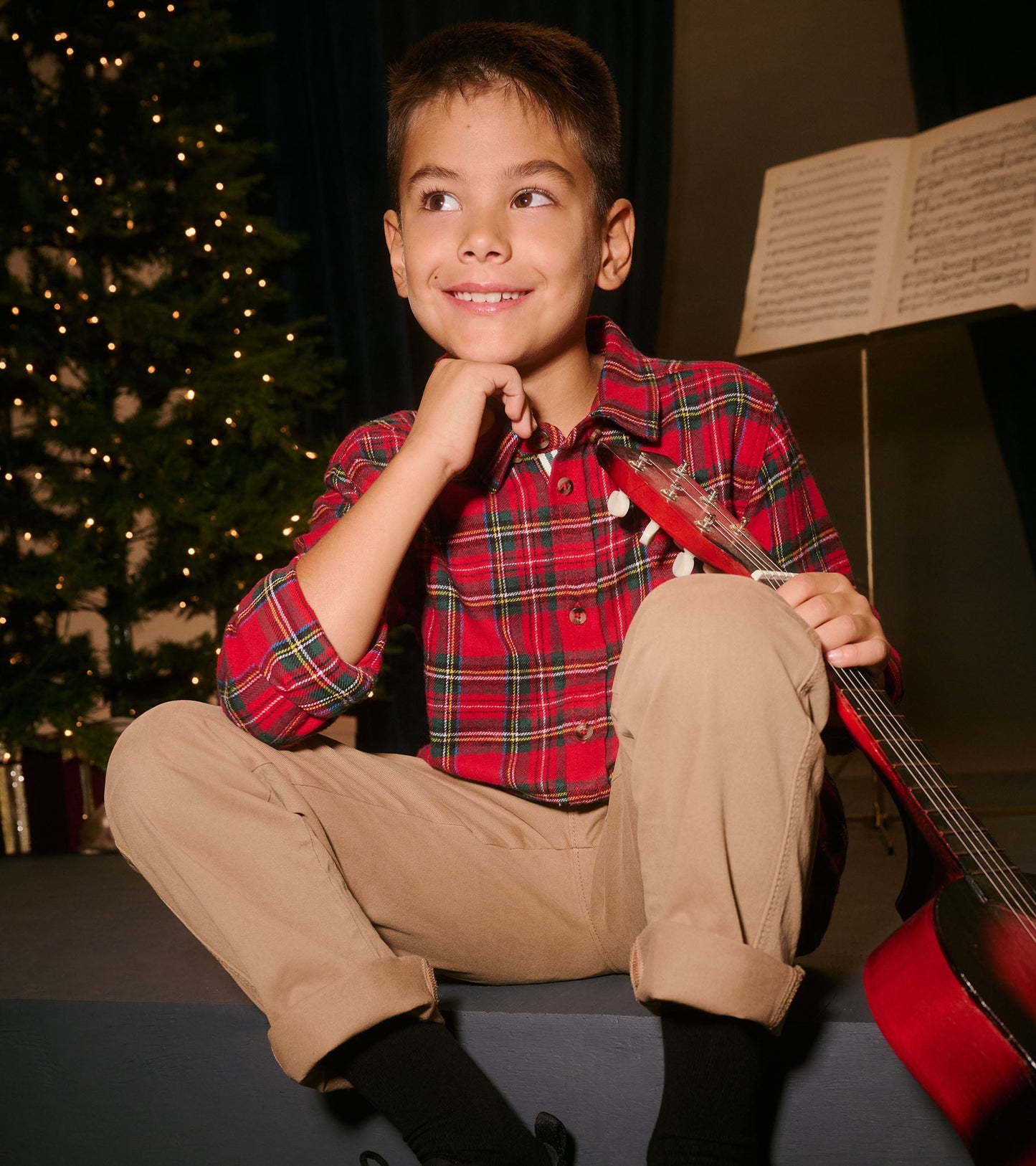 Red plaid button-down shirt worn by a boy, paired with khaki pants, sitting with a guitar in a festive setting.