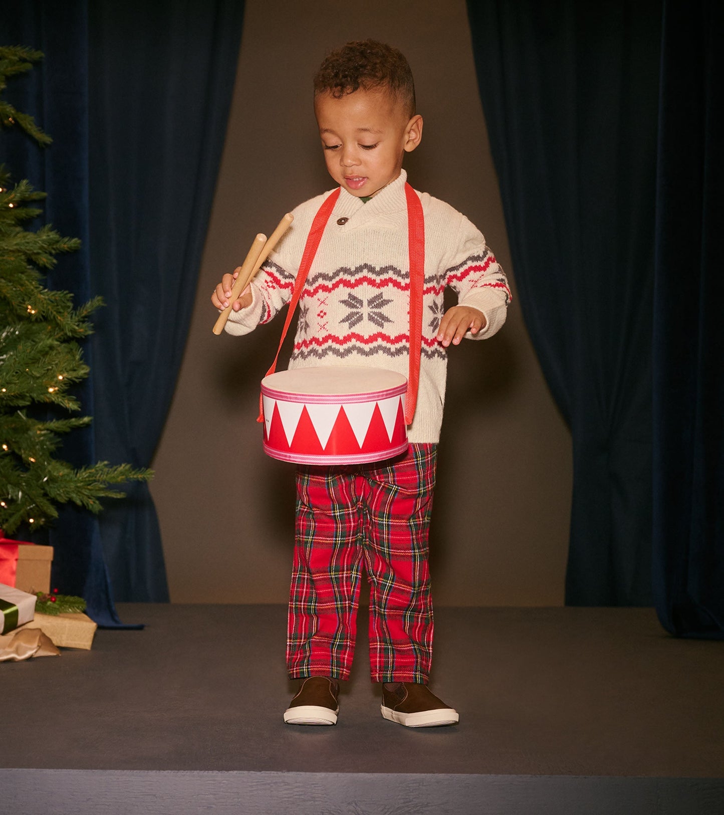 Red plaid straight leg pants for babies and toddlers, styled with a cozy sweater and drum, set against a festive backdrop.