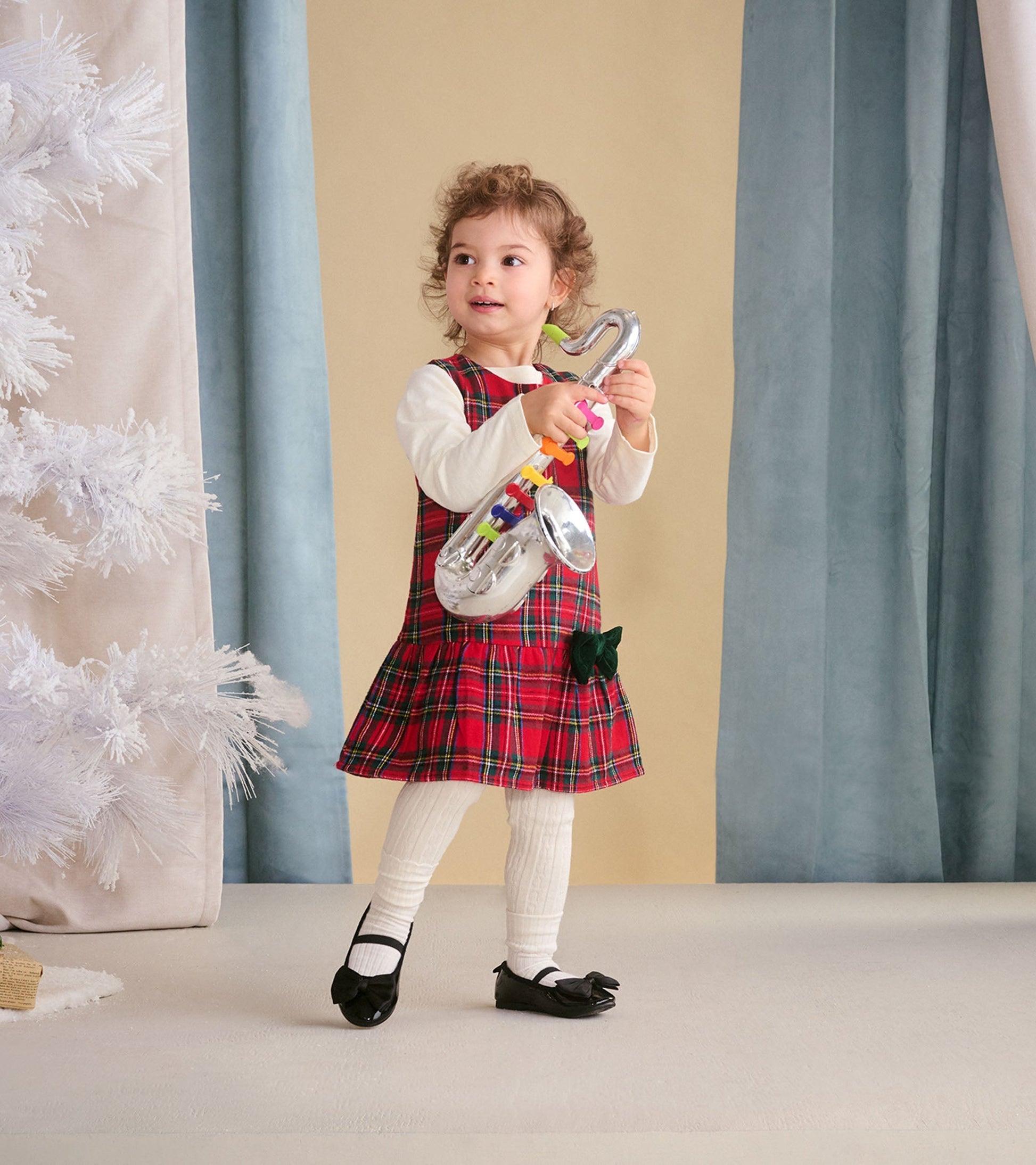 Red plaid pinafore dress with a flared skirt, worn over a long-sleeve white shirt, paired with white tights and black shoes.