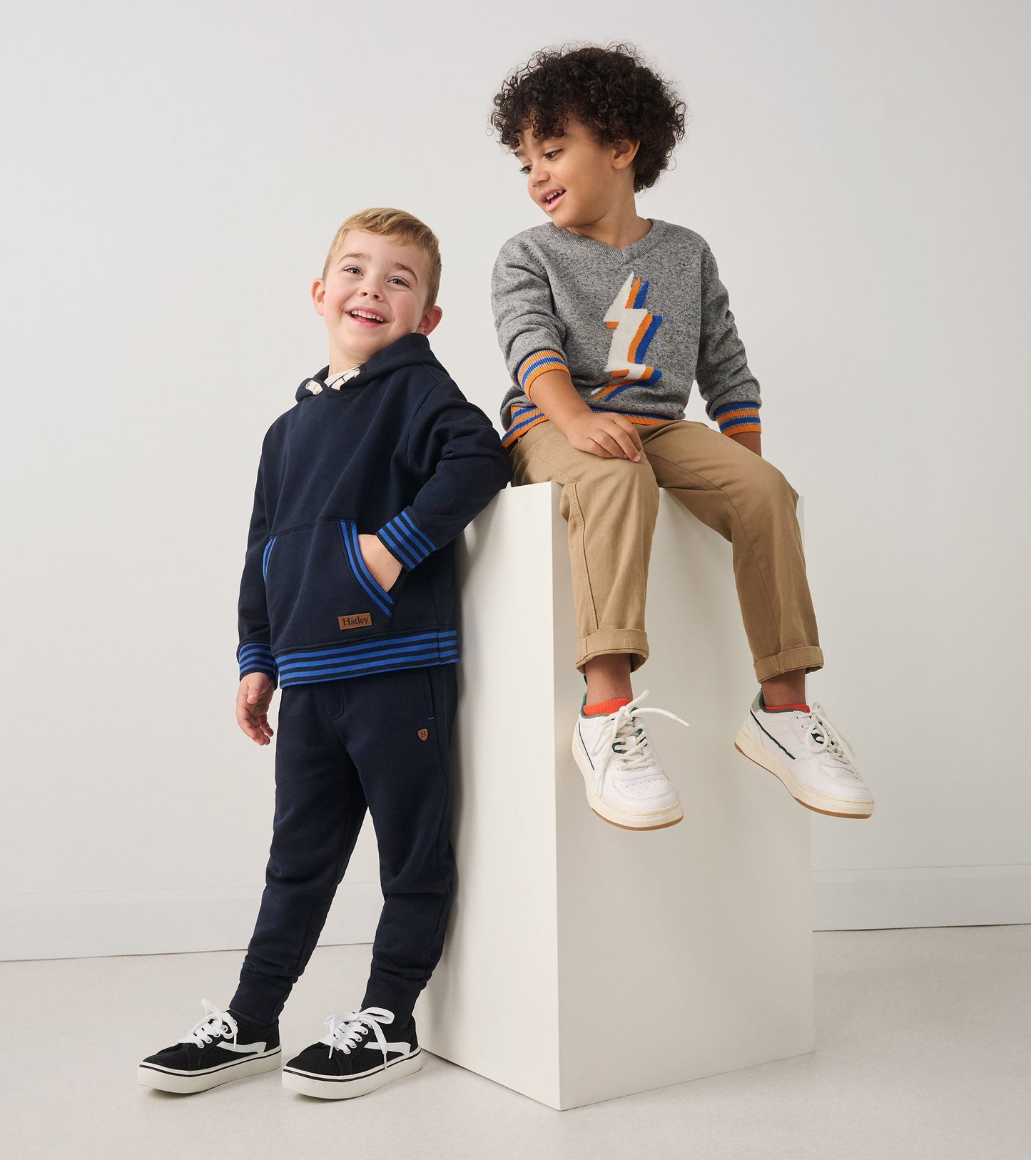 Navy pullover with blue stripes, worn by a boy, paired with matching pants and black sneakers. Another boy in a gray sweater…