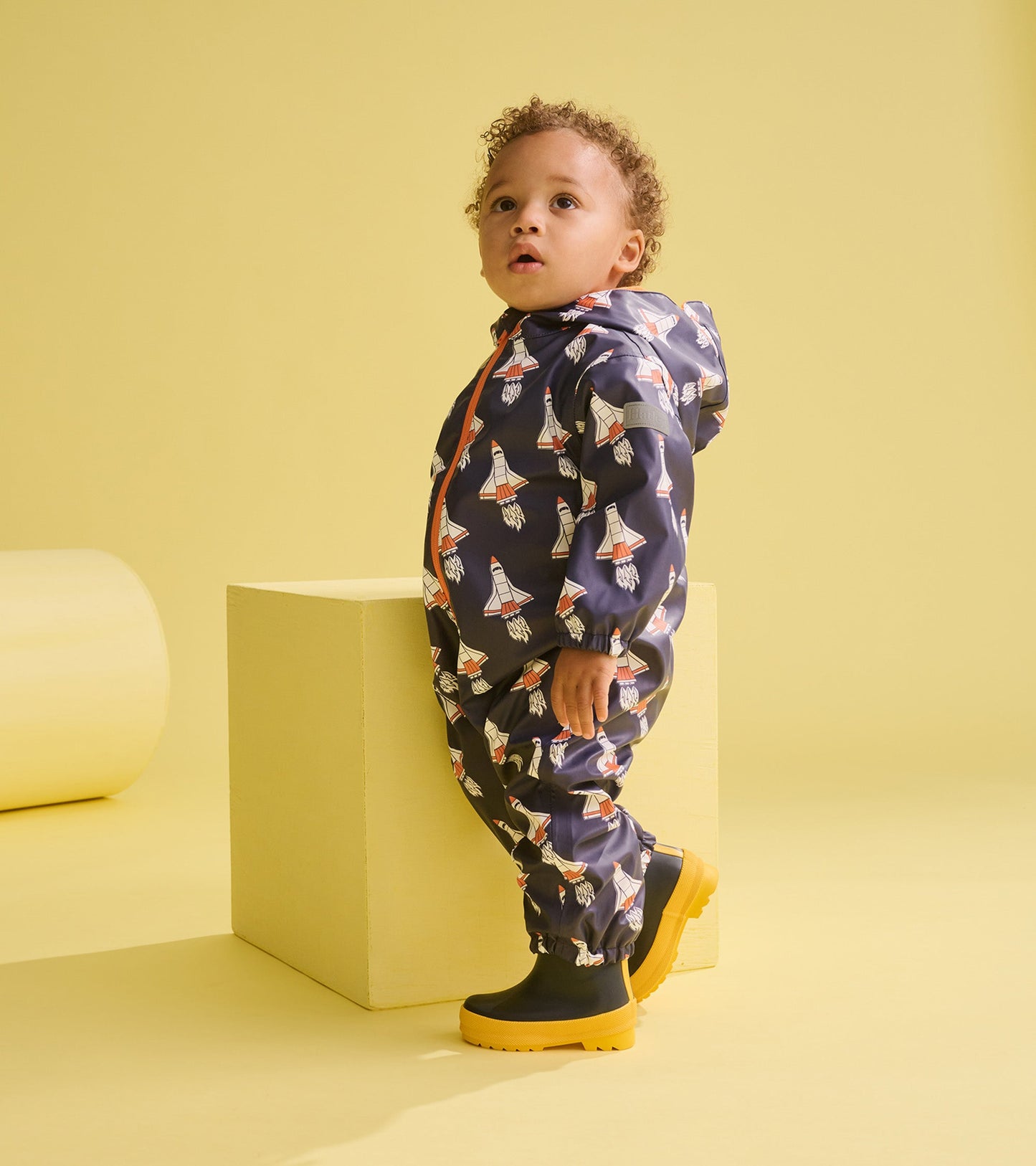 Navy rain suit for baby boys featuring colorful space shuttle prints, with a hood and elastic cuffs, paired with yellow boot…