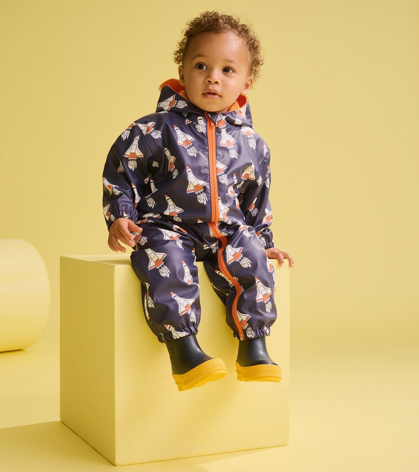 Navy rain suit for baby boys featuring colorful space shuttle prints, with an orange hood and elastic cuffs, worn with yello…