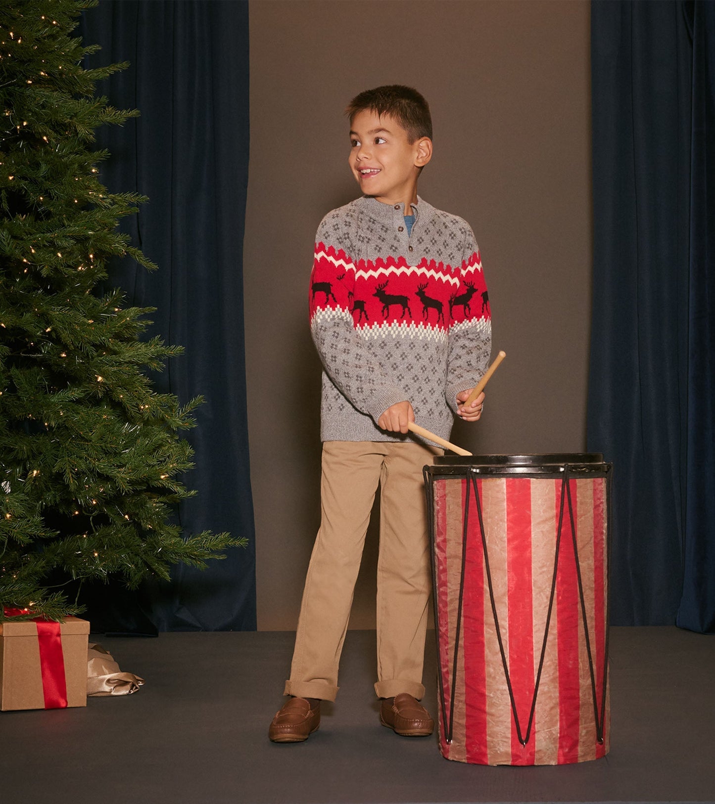 Gray mockneck sweater with red elk pattern, worn by a boy playing a drum, set against a festive backdrop.