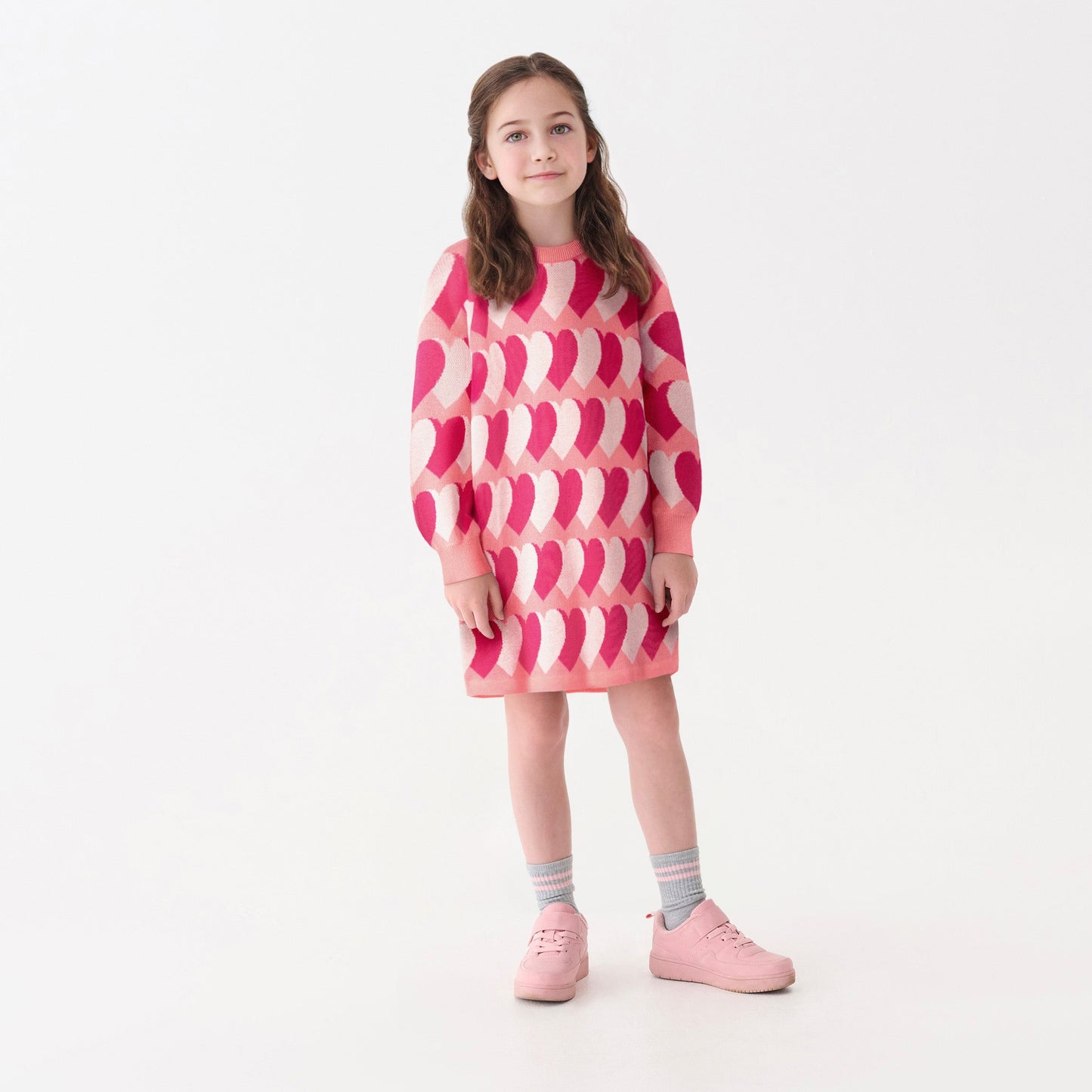 Pink sweater dress featuring a heart pattern, long sleeves, and a relaxed fit, worn with pink shoes and striped socks.