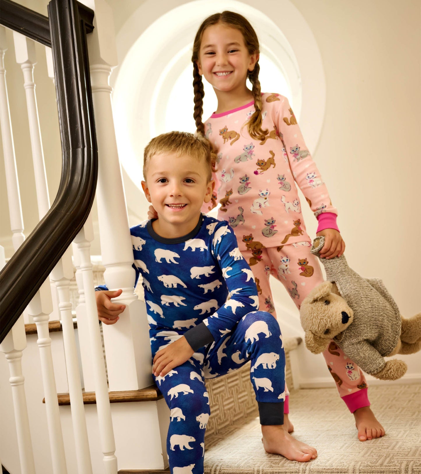 Cotton pyjama set featuring a blue bear print and a pink kitty pattern, worn by two children on stairs.