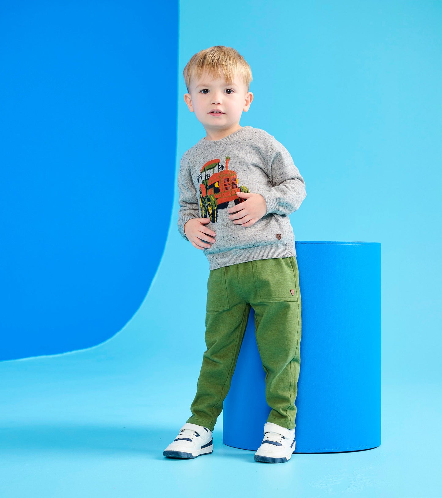 Gray V-neck sweater featuring a tractor graphic, paired with green pants, worn by a young boy against a blue background.