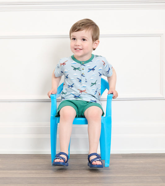 Light blue bamboo henley with airplane print, paired with green shorts, worn by a smiling toddler sitting on a blue chair.