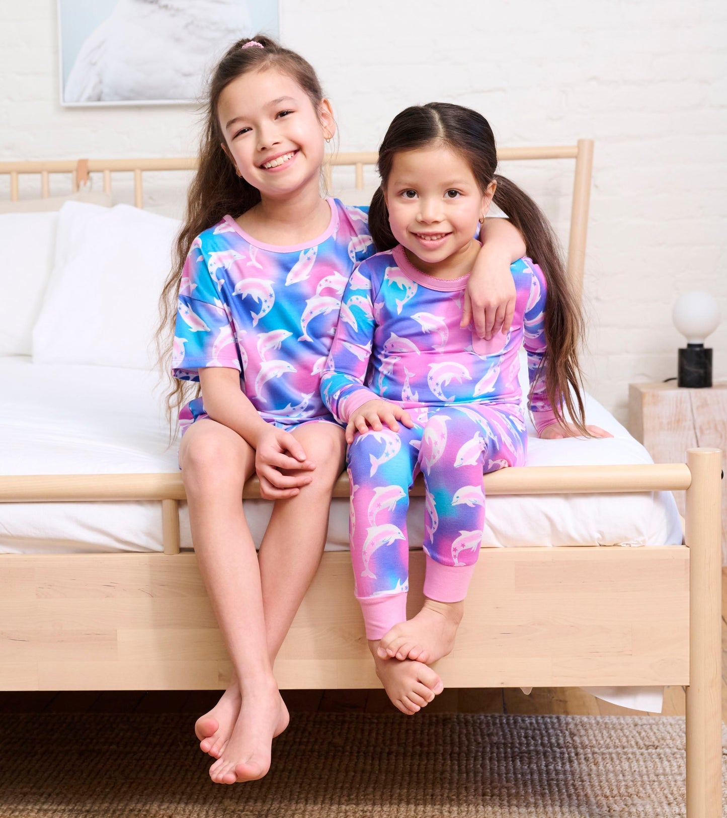 Girls in colorful dolphin-patterned bamboo pyjamas, sitting on a bed, smiling and embracing each other.