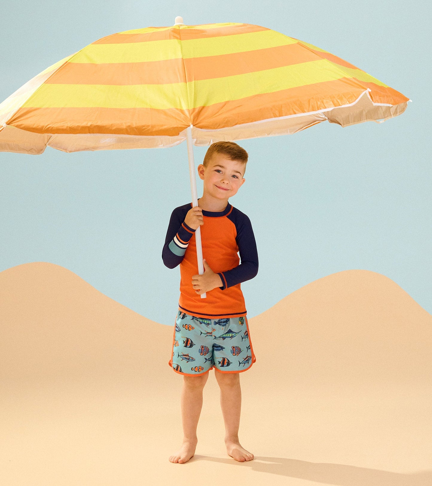 Boys' long sleeve rashguard in bright orange with navy sleeves, perfect for beachwear, paired with colorful swim shorts.