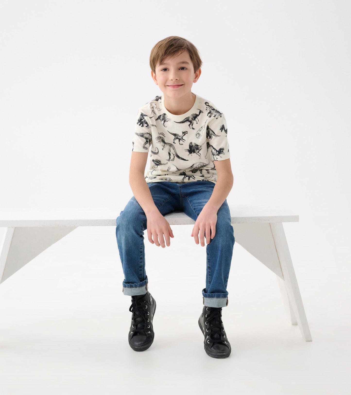 Cream graphic tee featuring black dinosaur prints, paired with blue jeans and black sneakers.