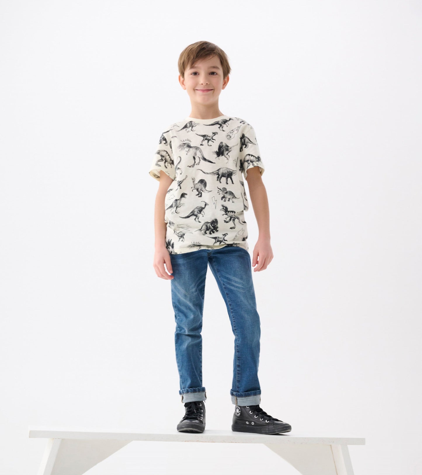 Cream graphic tee featuring black dinosaur prints, paired with blue jeans and black sneakers. Model standing on a white plat…