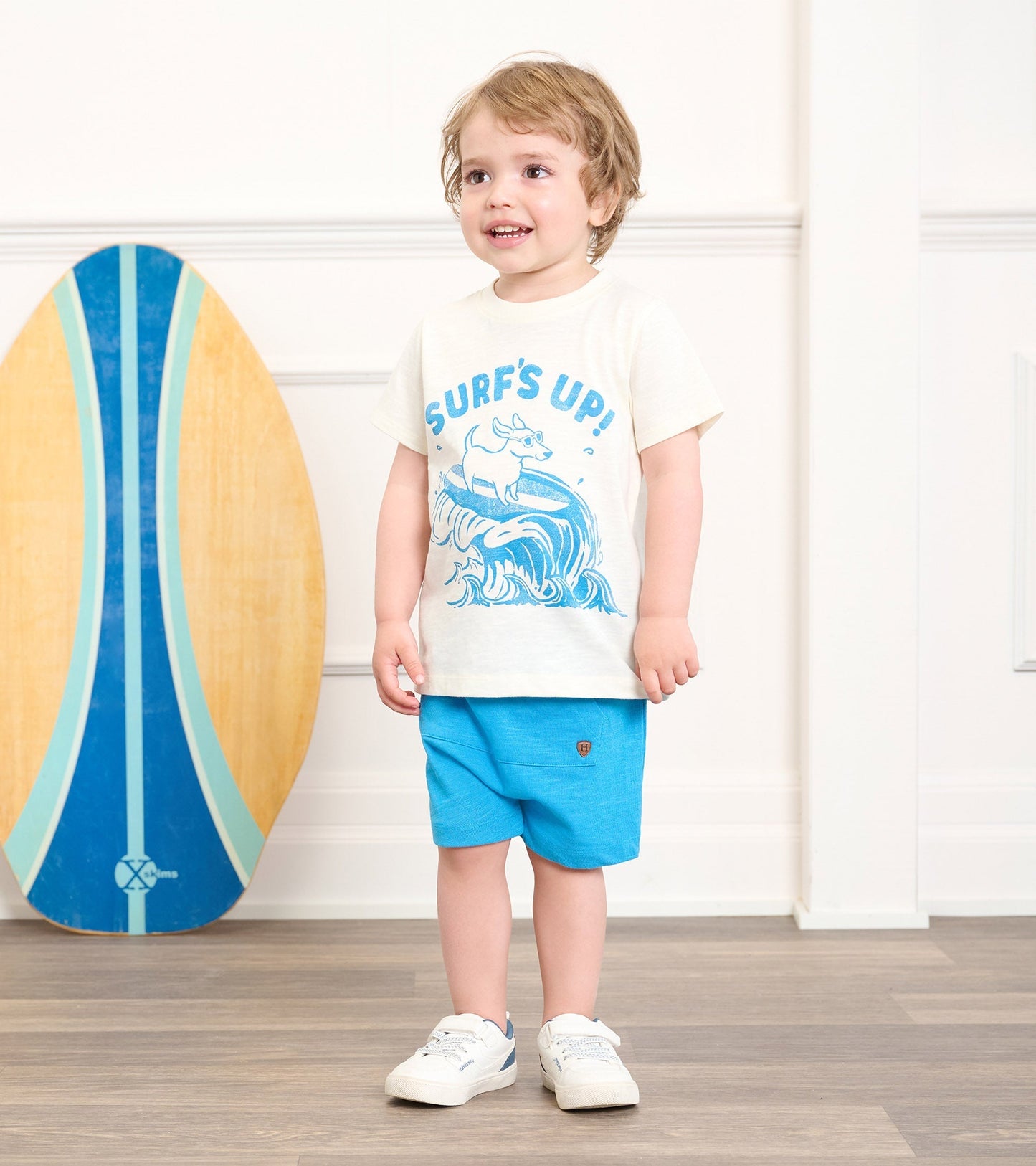 Light cream graphic tee with blue surf design, paired with bright blue shorts, worn by a smiling toddler.