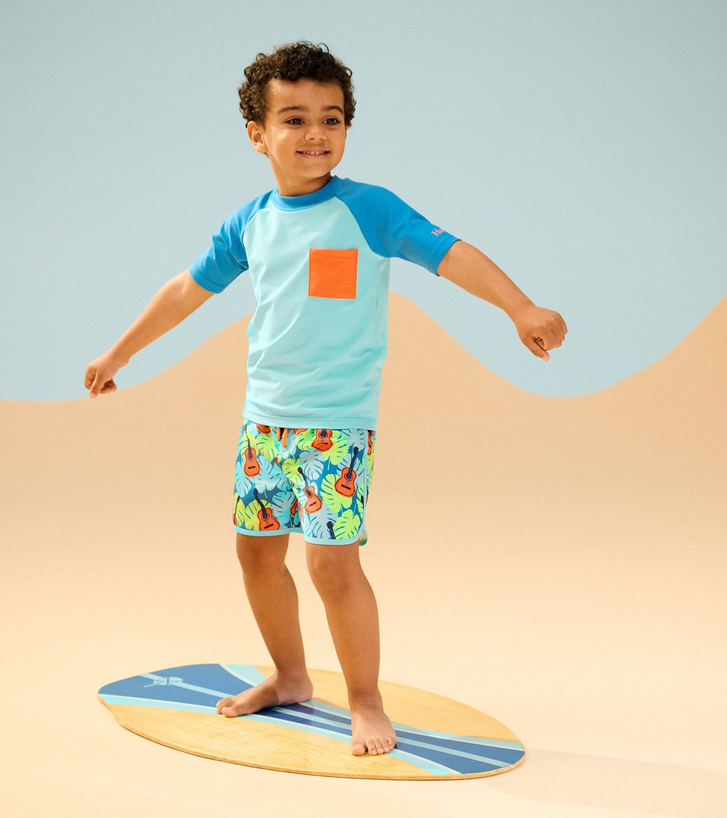 Light blue short sleeve rashguard with orange pocket, paired with colorful patterned swim shorts, worn by a smiling boy.
