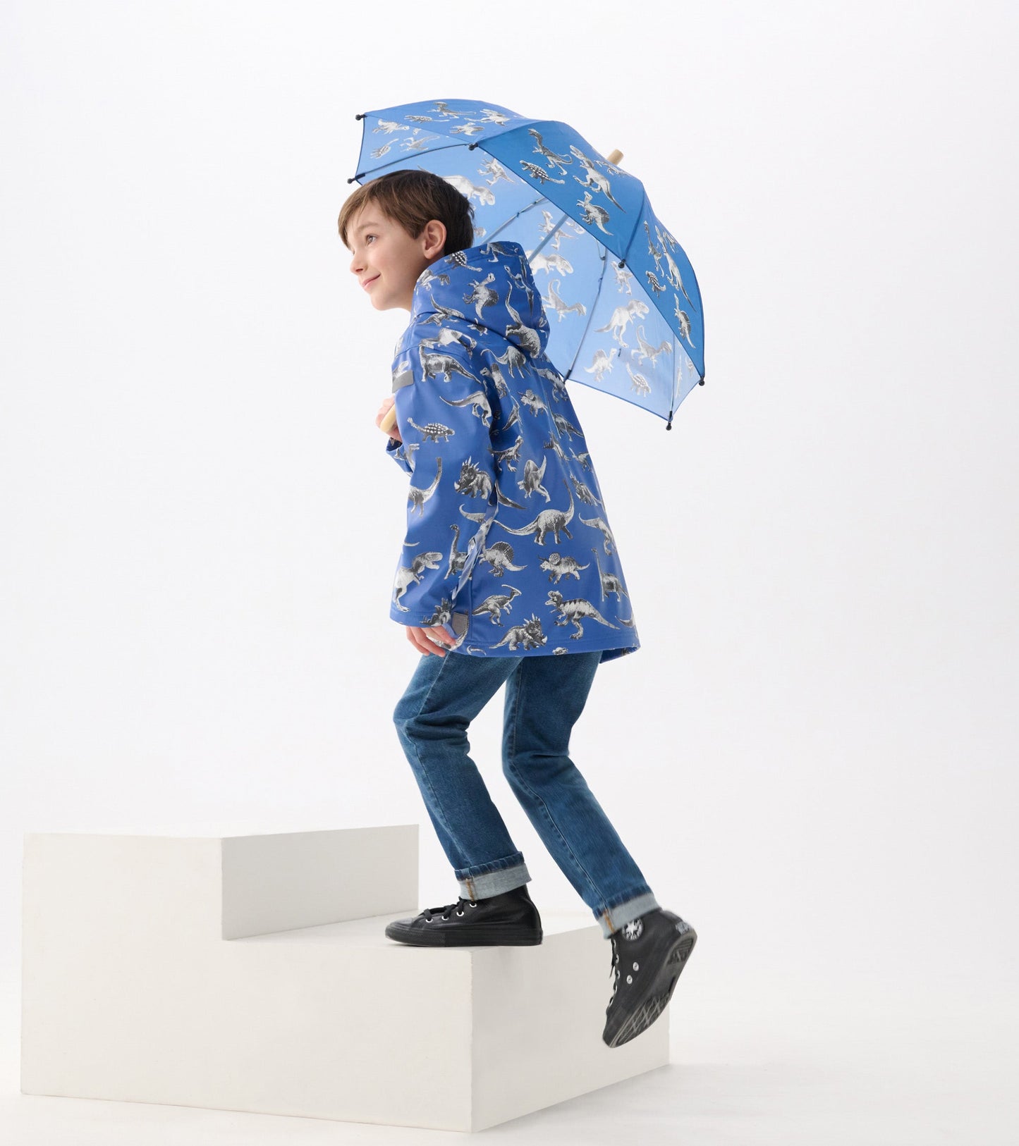 Blue umbrella featuring dinosaur patterns, held by a child wearing a matching raincoat and jeans, stepping up a white platfo…