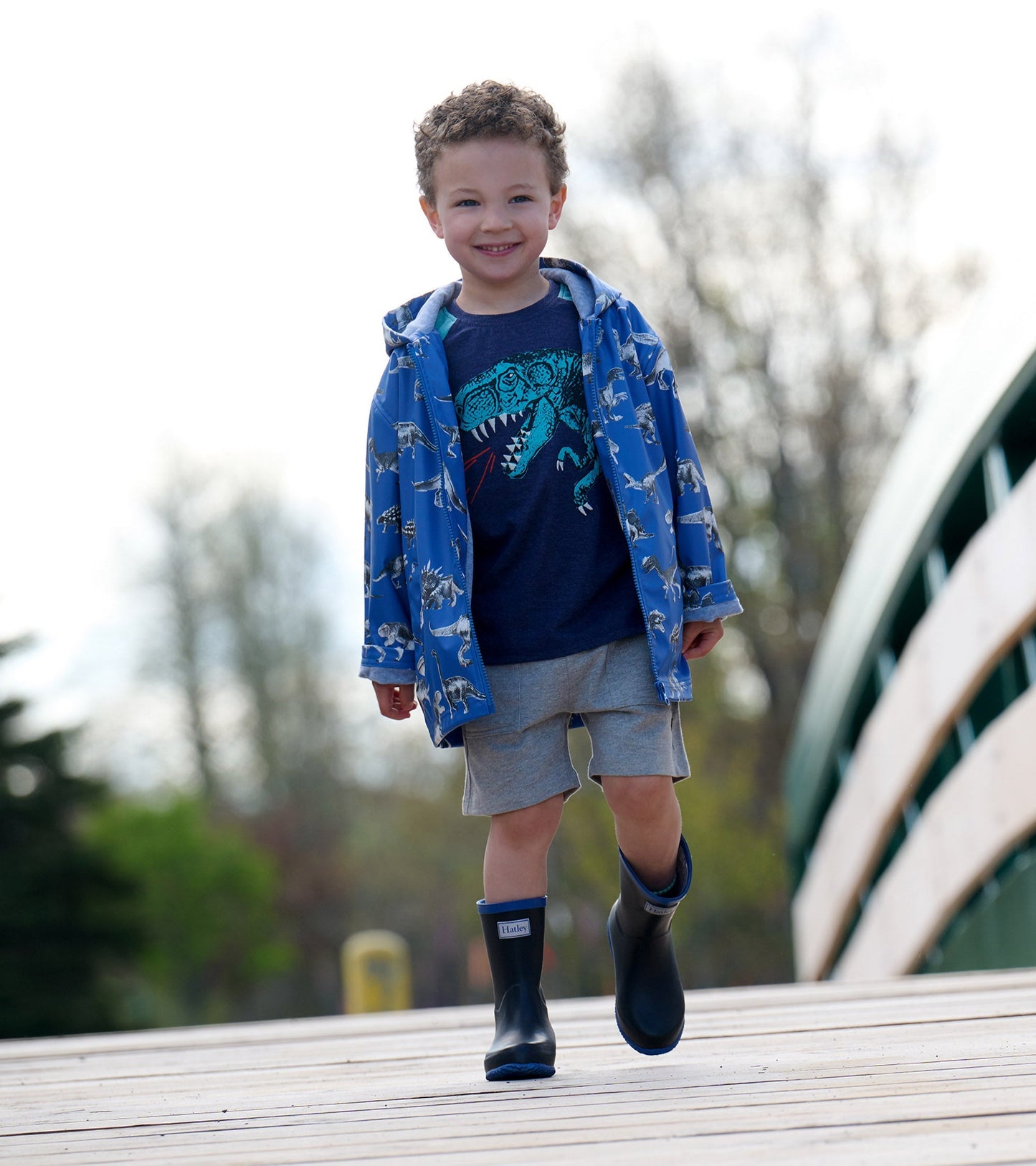 Blue zip-up raincoat with dinosaur print, worn by a smiling boy, paired with a dark t-shirt and gray shorts.
