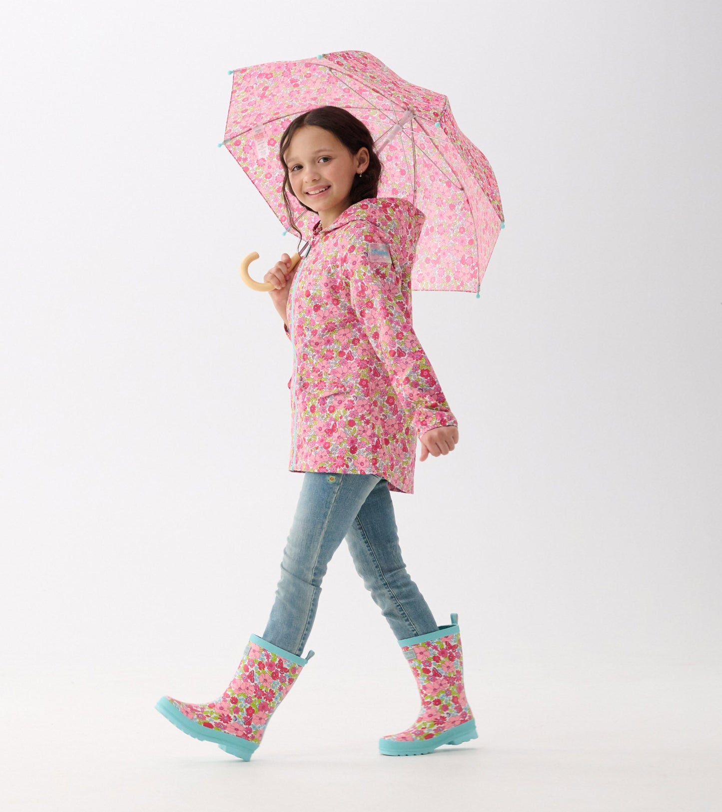 Pink umbrella with colorful flower pattern, held by a girl in a matching raincoat and boots, walking confidently.