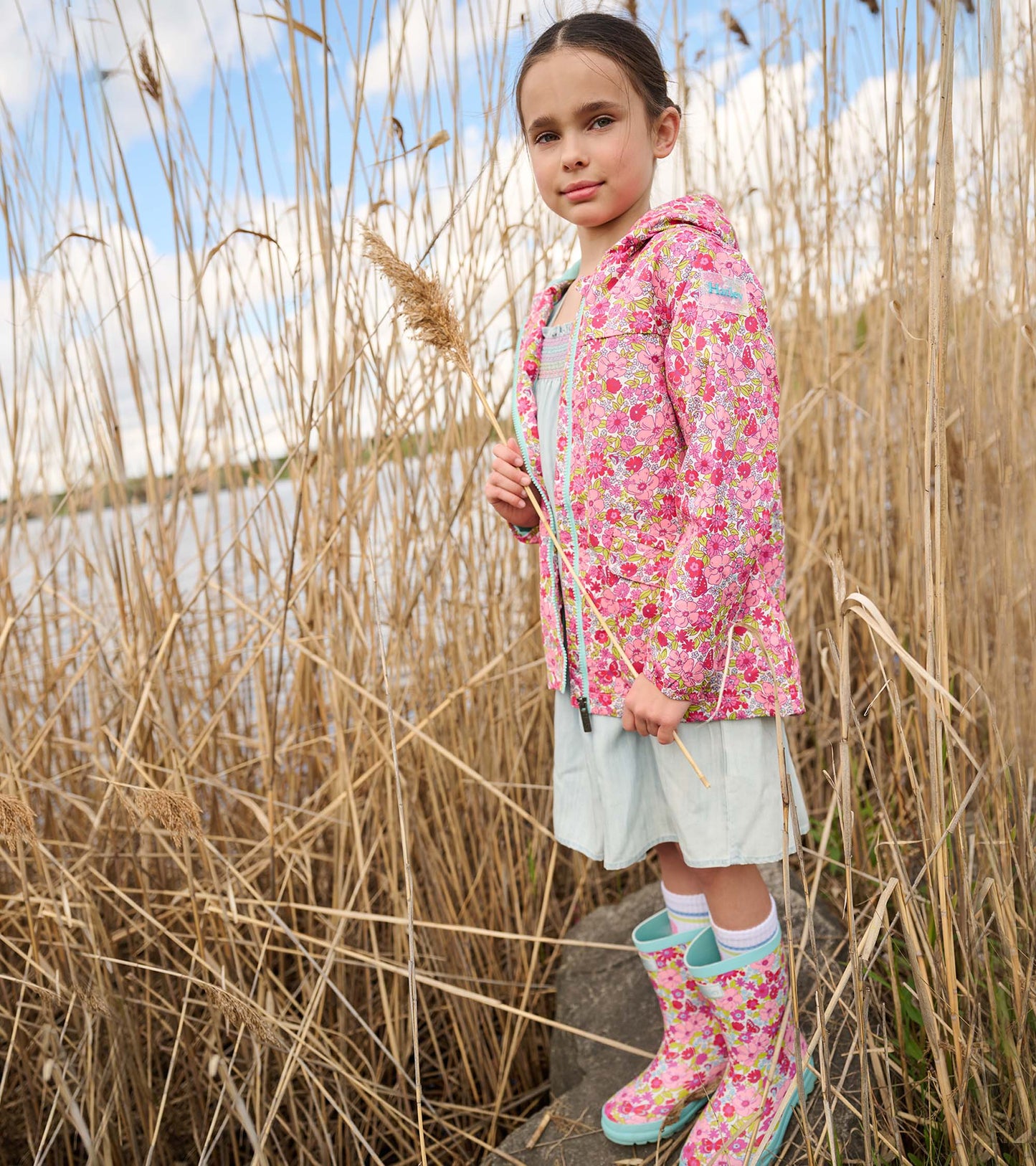 Lightweight pink raincoat with a floral pattern, worn by a girl standing in tall grass near water.