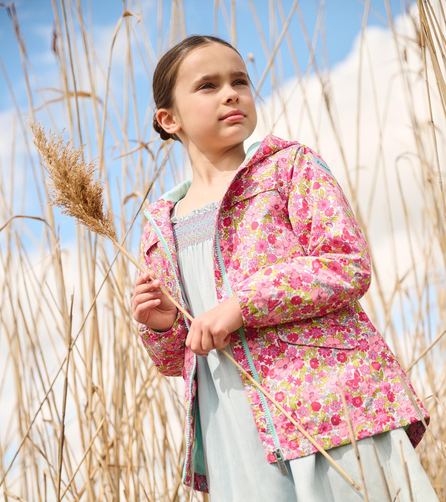 Lightweight pink raincoat with a colorful floral pattern, worn by a girl holding a dried plant in a natural setting.