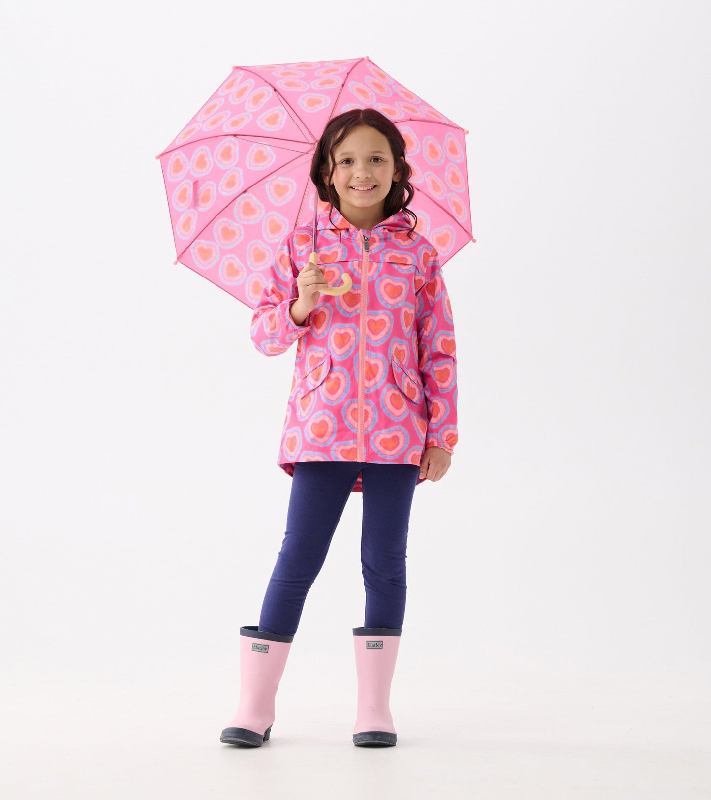 Pink umbrella with glowing heart patterns, held by a girl wearing a colorful raincoat and pink rain boots.