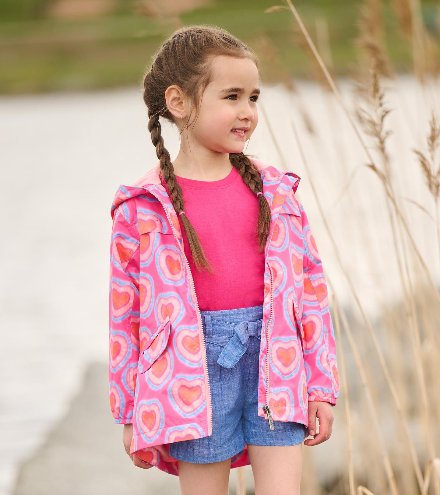 Lightweight zip-up raincoat in pink with colorful heart patterns, worn by a girl with braids, paired with a pink top and blu…
