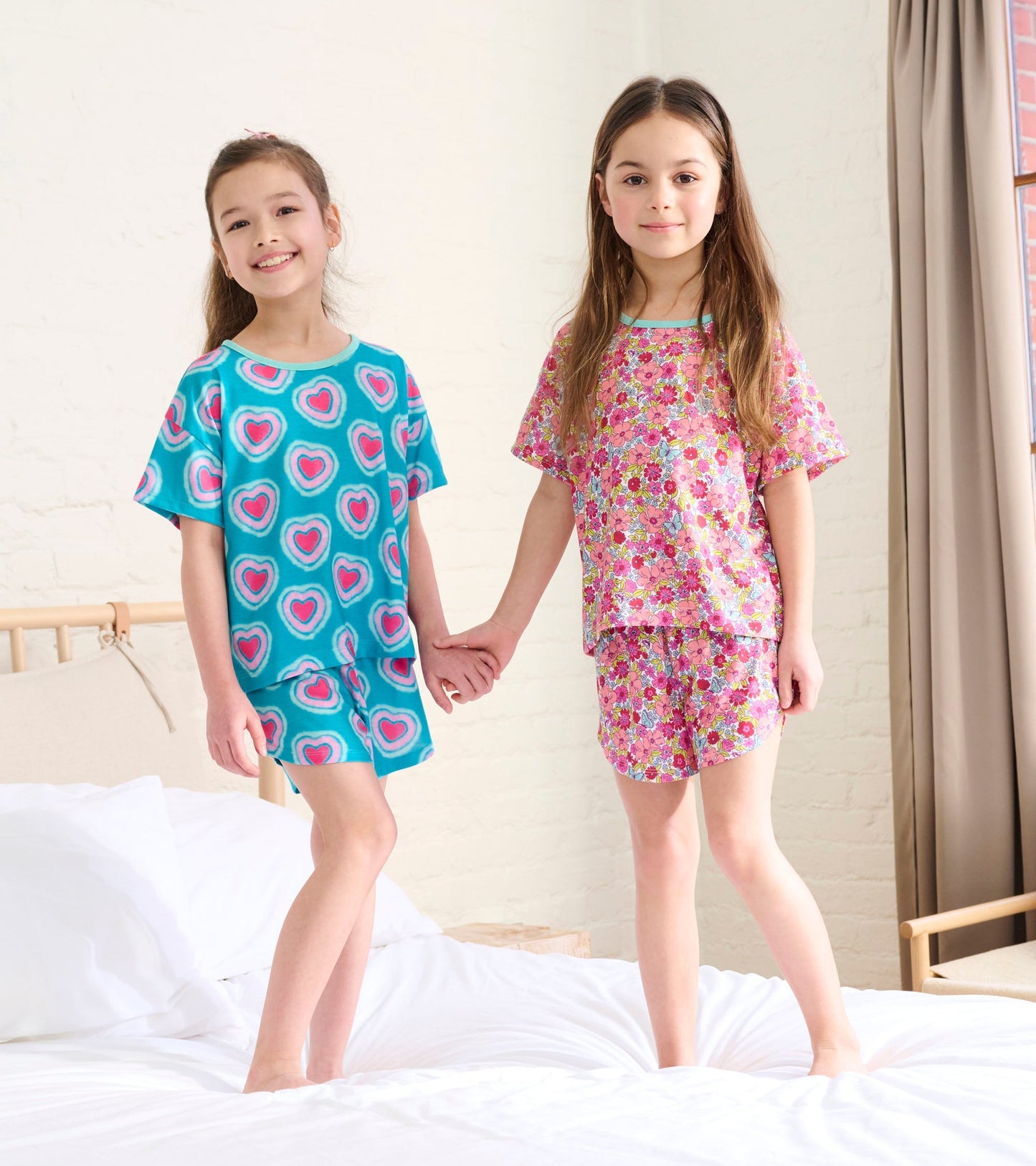 Two girls holding hands in colorful heart and floral patterned pyjama sets, featuring short sleeves and shorts.