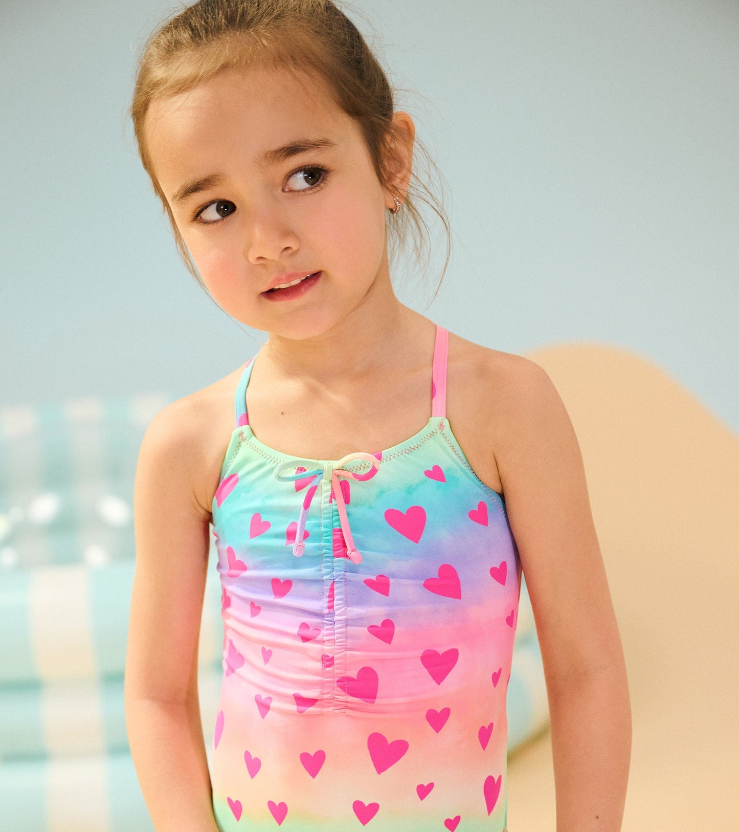 Pastel-colored swimsuit with ruched front and pink heart pattern, featuring adjustable straps and a tie detail.