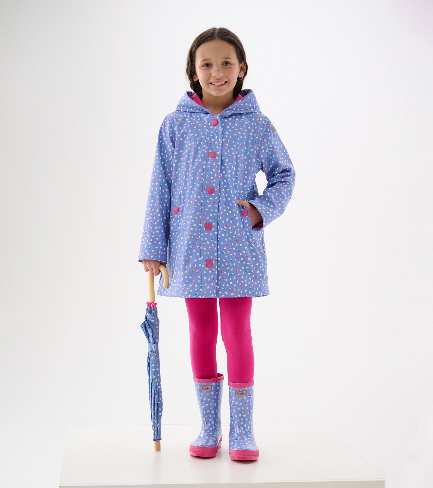 Light blue button-up raincoat with colorful dots, hood, and pink buttons, paired with pink leggings and matching rain boots.