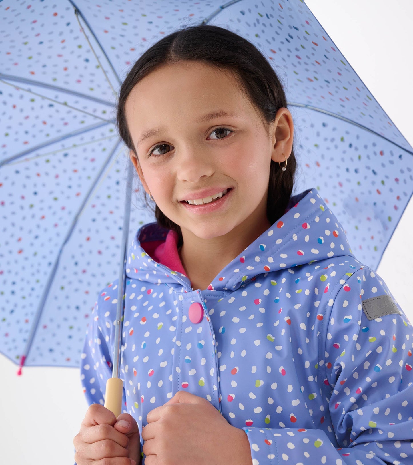 Light blue raincoat with colorful dot pattern, featuring a hood and button closure, held by a smiling girl with an umbrella.