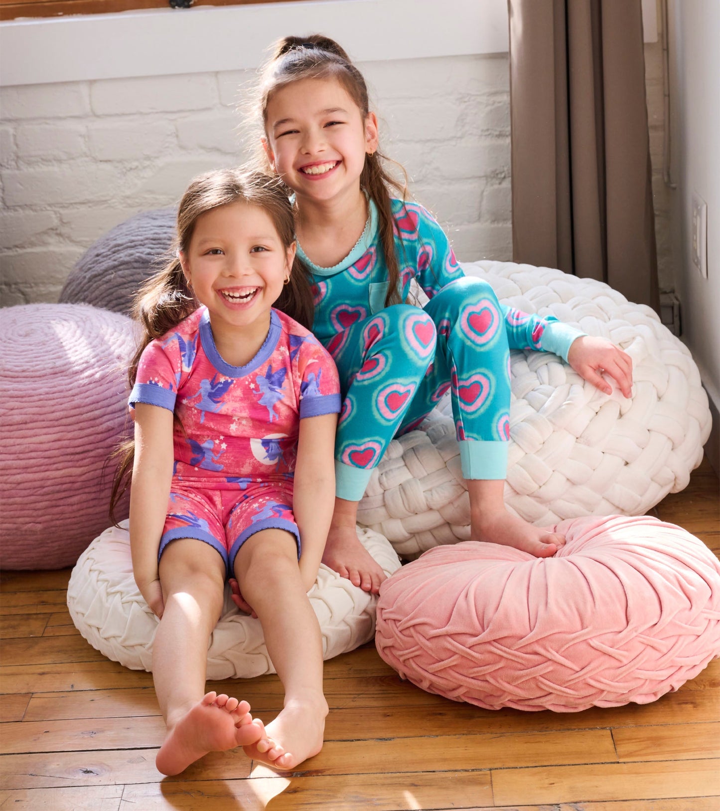 Two girls sit on colorful cushions, wearing a pink fairy-patterned short pyjama set and a teal heart-patterned long pyjama s…