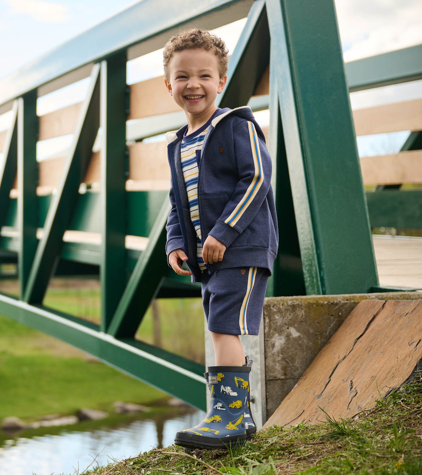 Patriot blue pull-on shorts with side stripes, worn by a smiling boy standing near a bridge and wearing rain boots.
