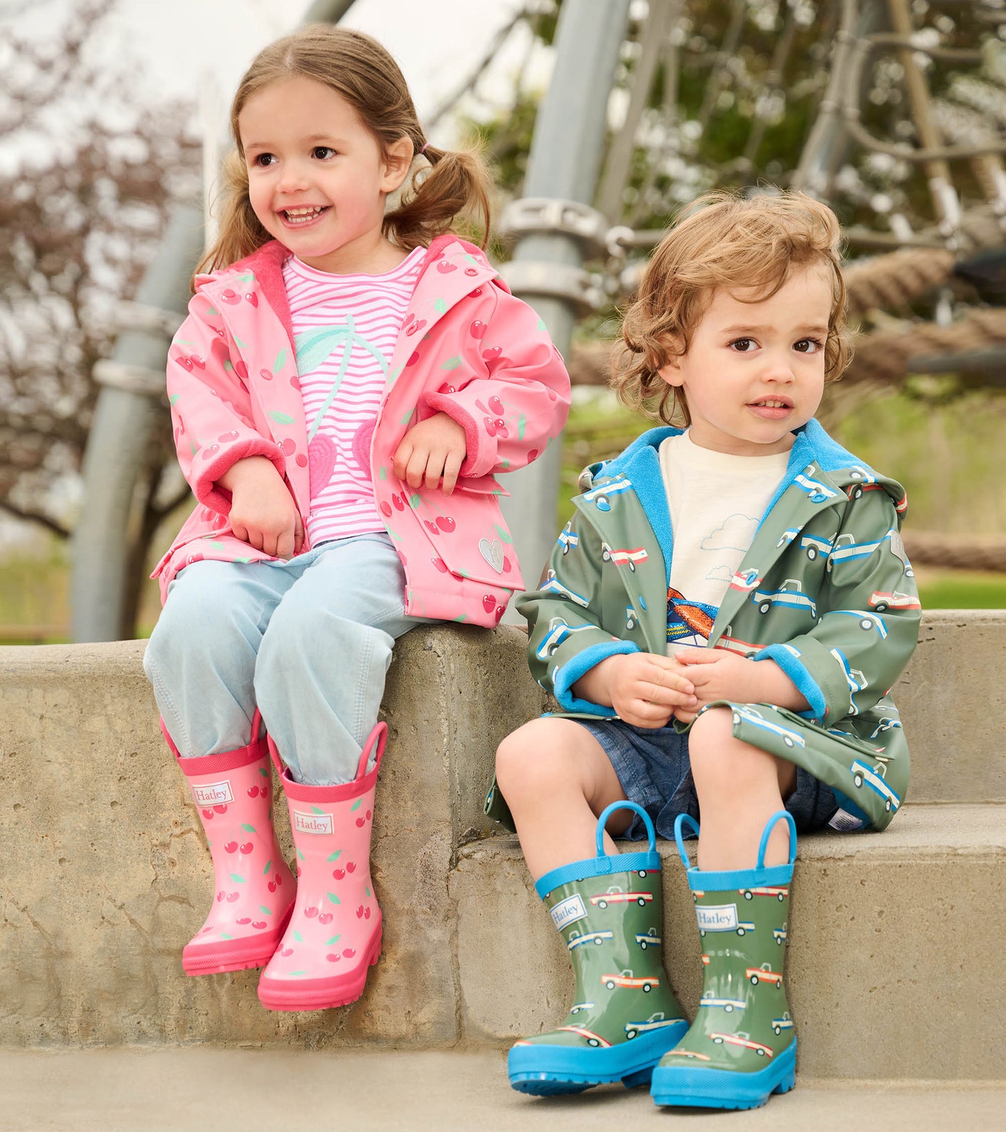 Pink raincoat with cherry pattern on a girl, and a green raincoat with car pattern on a boy, both wearing colorful boots.