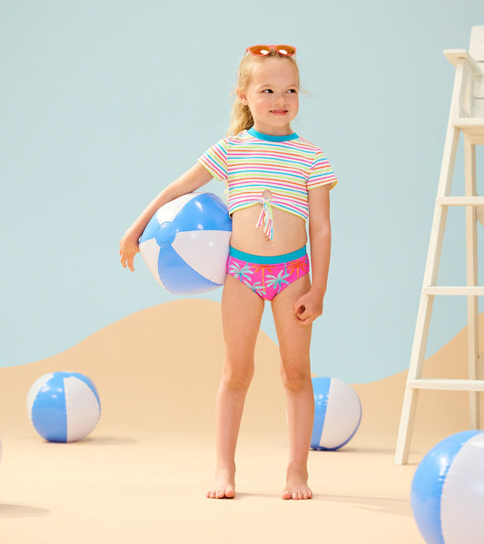 Colorful striped short sleeve tankini with palm print bottoms, perfect for beach play, worn by a smiling girl holding a beac…