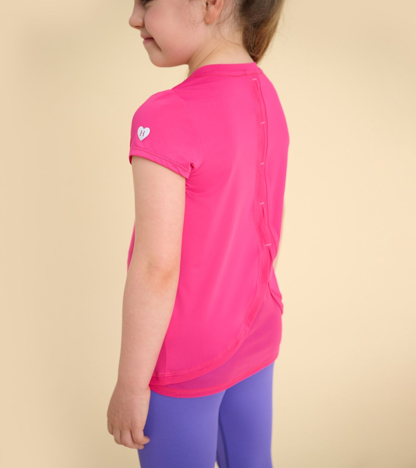 Pink short-sleeve t-shirt with mesh trim and button detail at the back, worn by a girl with purple leggings.